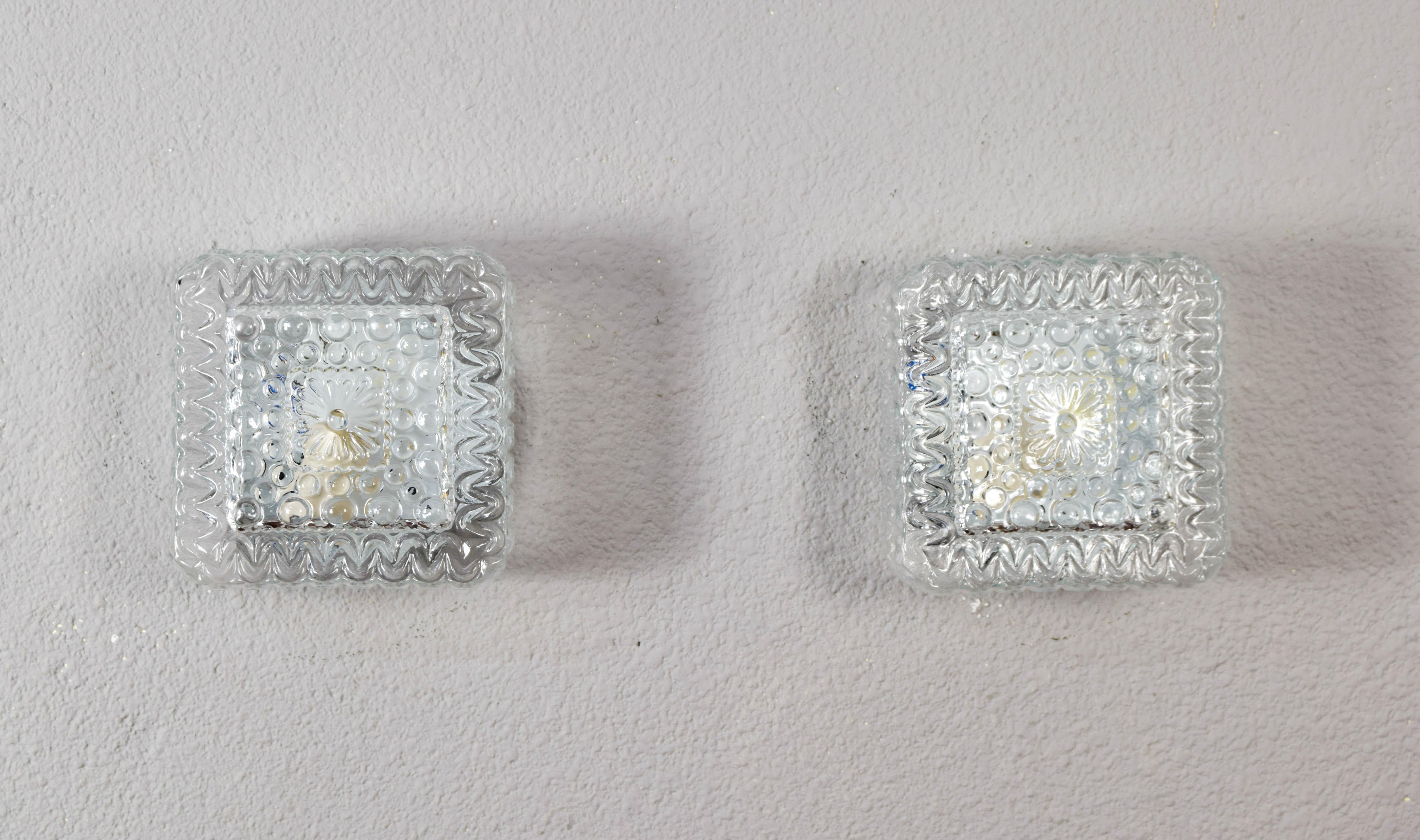 Beautiful set consisting of two wall lights or flush mount lamps of Germany origin manufactured in the 1960s.
Square in shape and made of transparent cut glass, they consist of an E27 light bulb socket.
The shapes of the glass create a very pleasant