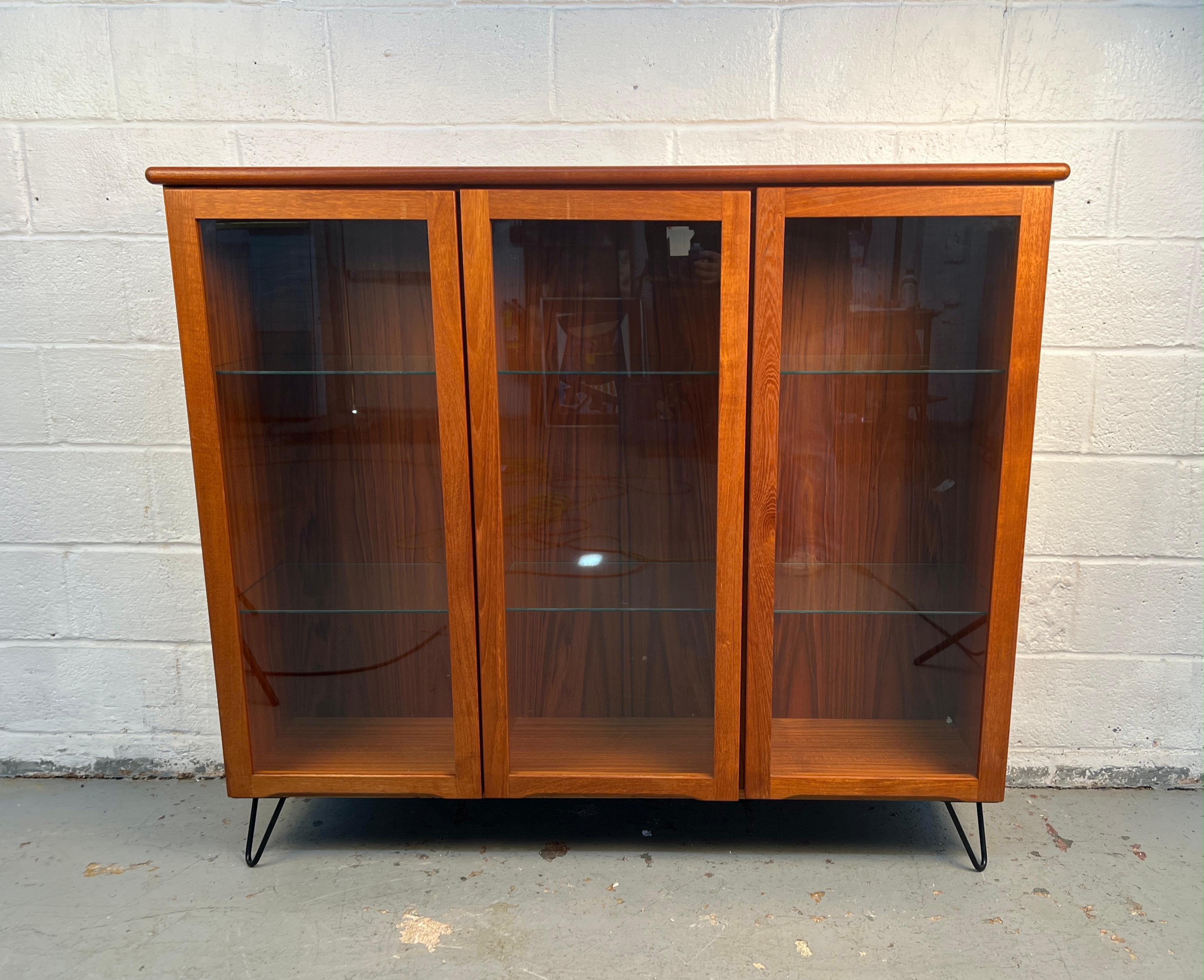 Una libreria espositiva moderna di metà secolo realizzata in caldo teak e dotata di eleganti ante in vetro. Il mobile offre un mix equilibrato di visibilità aperta e spazio chiuso, rendendolo ideale per esporre libri, oggetti da collezione o