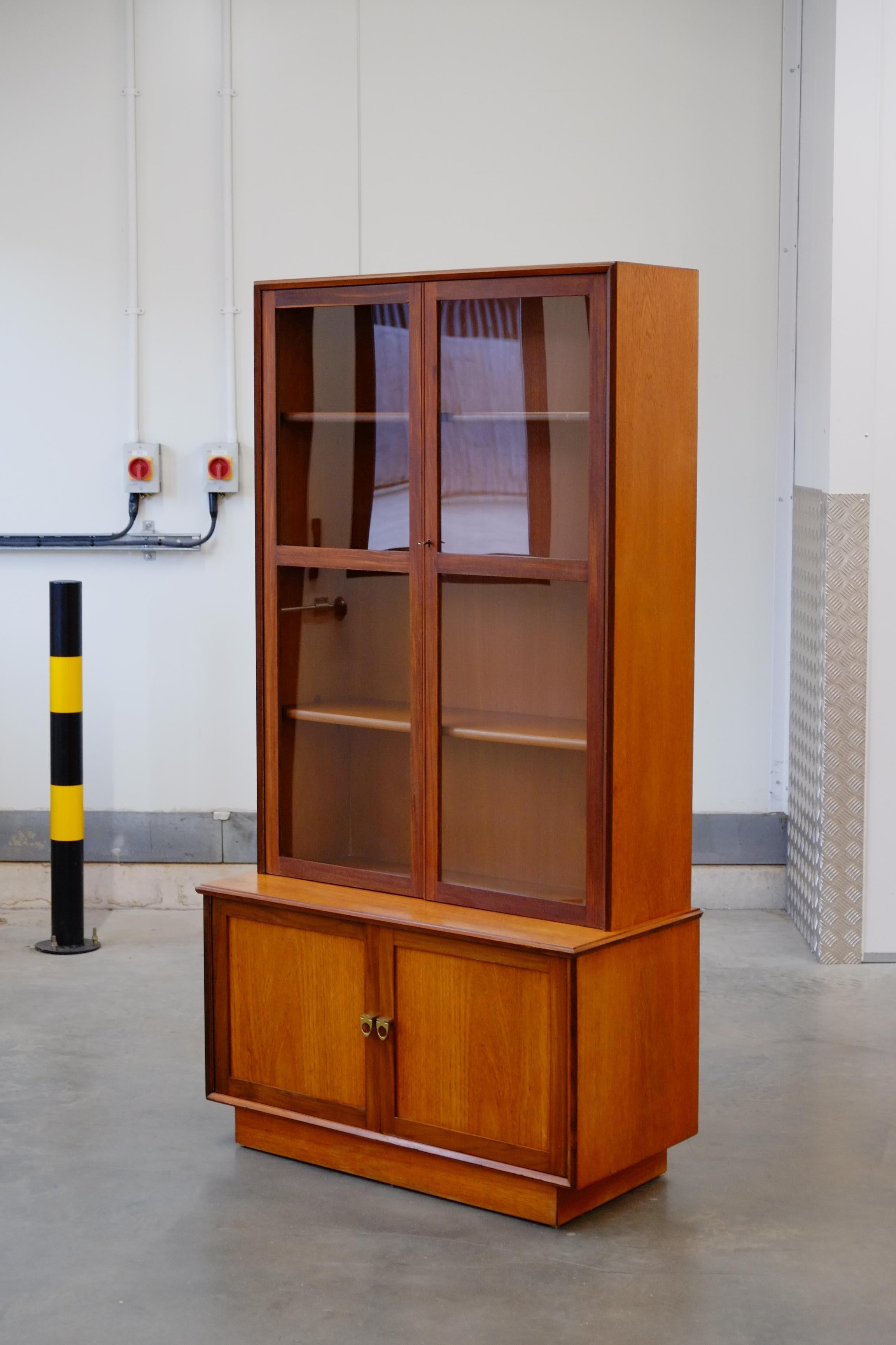 A beautiful tall slim glazed bookcase from the 1960s. This Mid-century modern cabinet is made in a lovely stripy Teak wood with a gorgeous warm color. The cabinet has elegant design features such as the cubic plinth base, angled edges, which add a