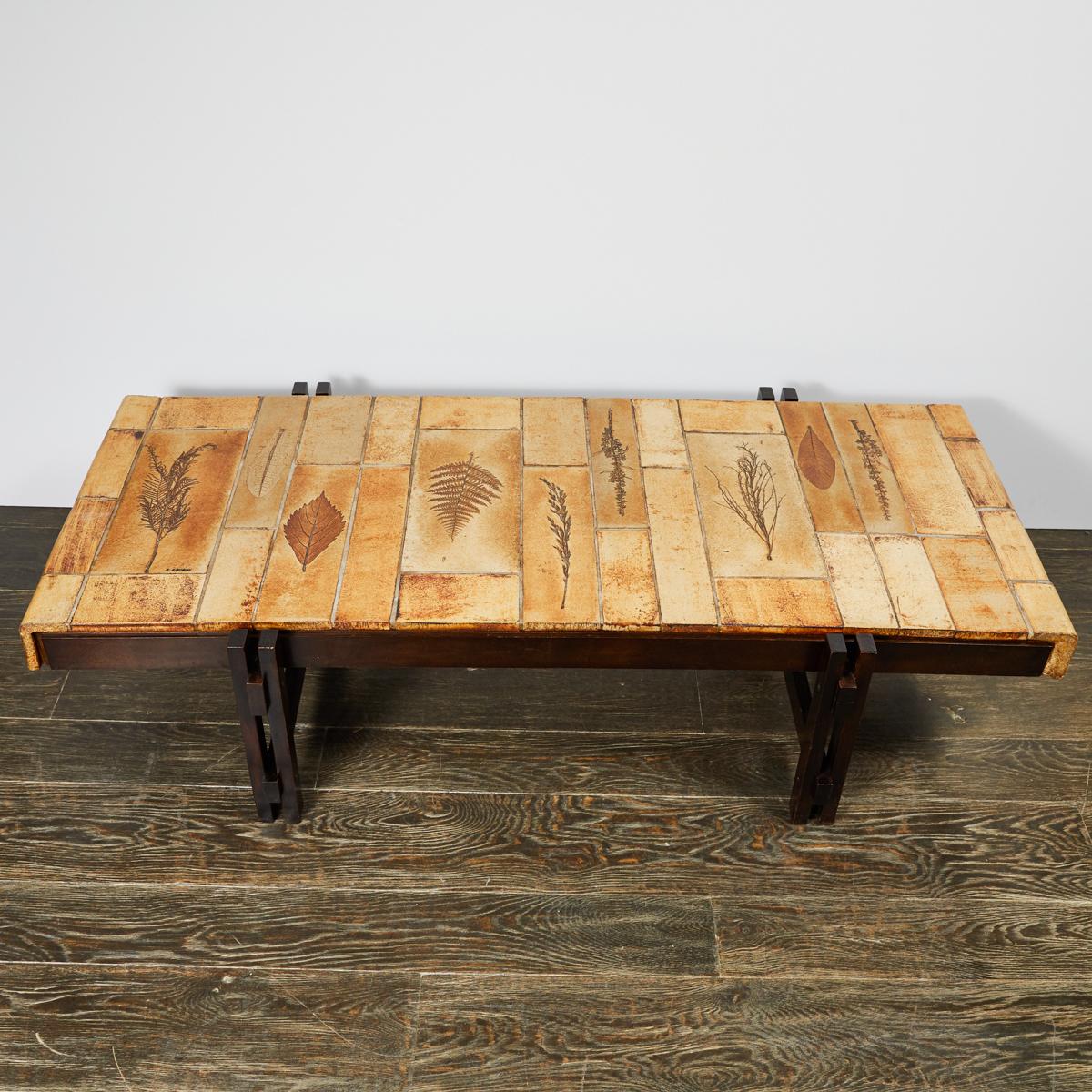 Roger Capron signed tile-top coffee table with carved walnut wood base. The tiles have been finished with a warm, golden glaze, and feature a motif of various leaf impressions. An organic yet industrial piece of French mid-century design. 

France,