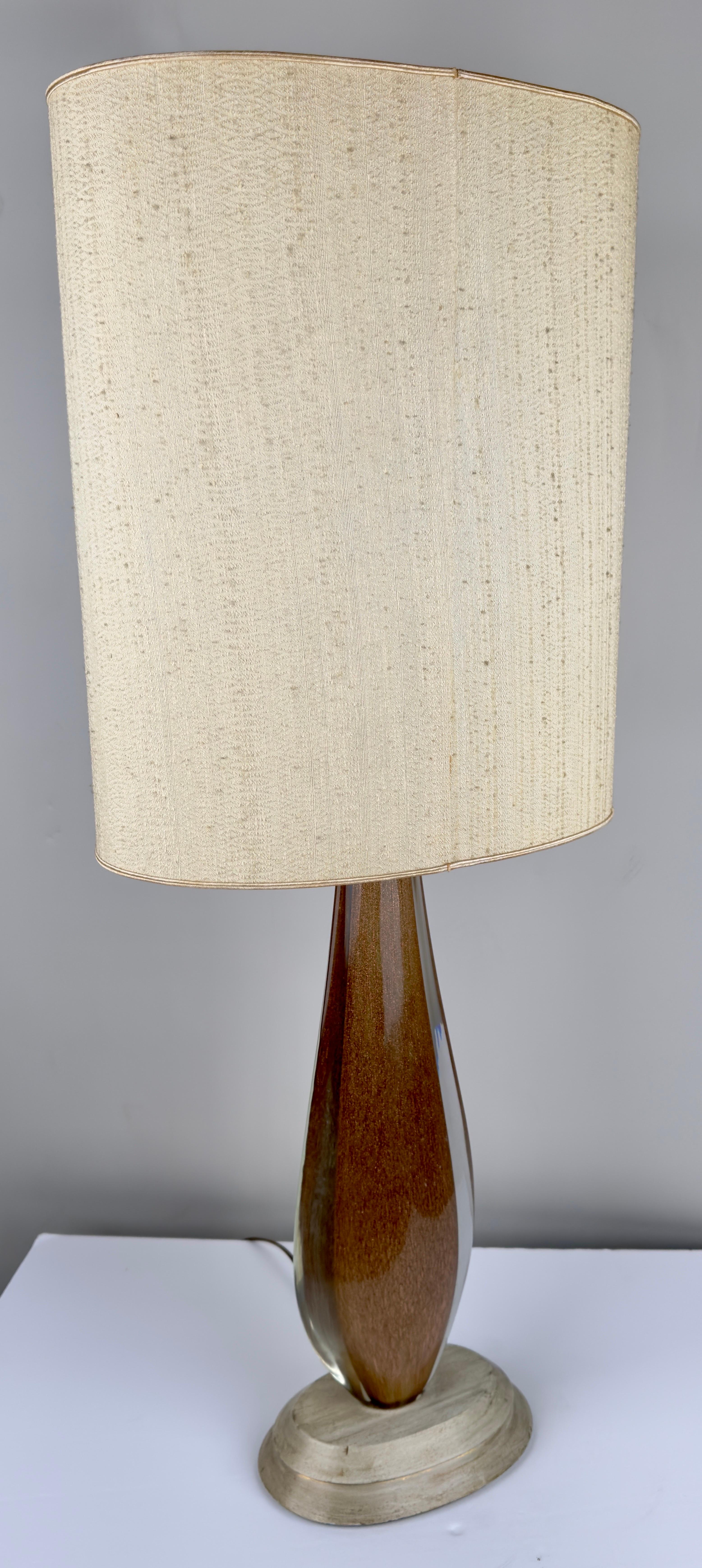  A  Mid Century Modern table lamp. Crafted from art glass adorned with golden grains that visually seem to dance within the glass, the lamp exudes a sense of timeless elegance and allure.

The curvaceous body of the lamp, resplendent in its opulence