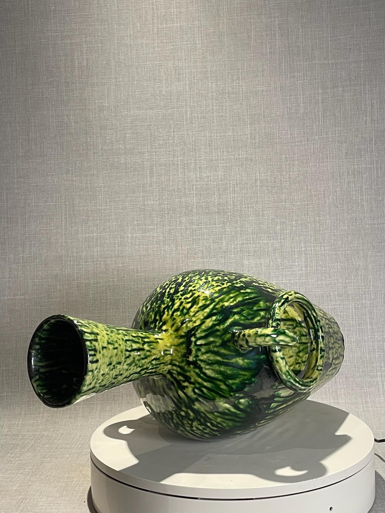 Ce vase en terre cuite du milieu du siècle dernier attire l'attention avec sa glaçure verte et noire et ses poignées sculpturales à double anneau. La pièce présente une silhouette haute, inspirée des amphores, avec un col évasé et des épaules