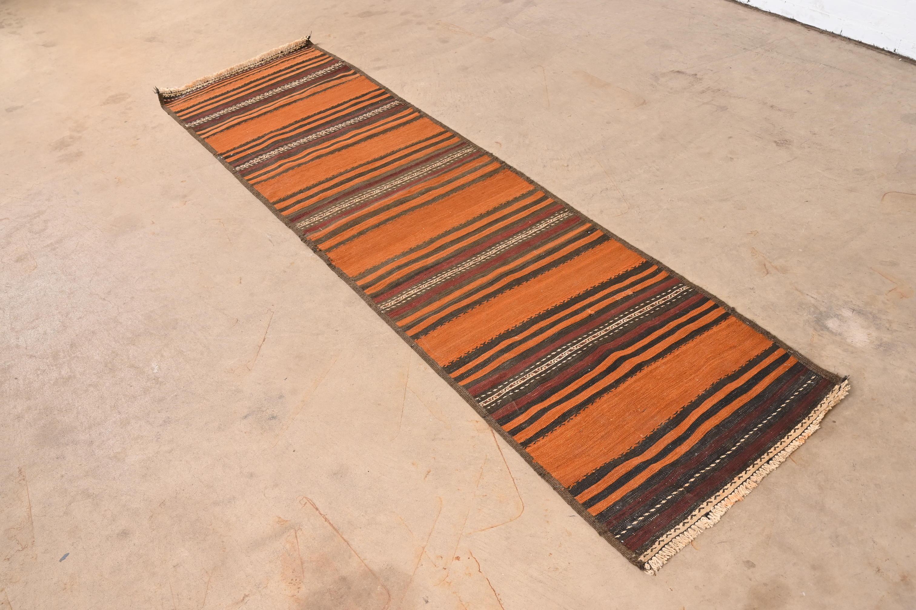 A gorgeous Mid-Century Modern hand-woven Afghan Kilim flat weave runner rug.

Mid-20th century.

Beautiful striped design, with predominant colors in orange, maroon, black, and ivory.

Measures: 26.5