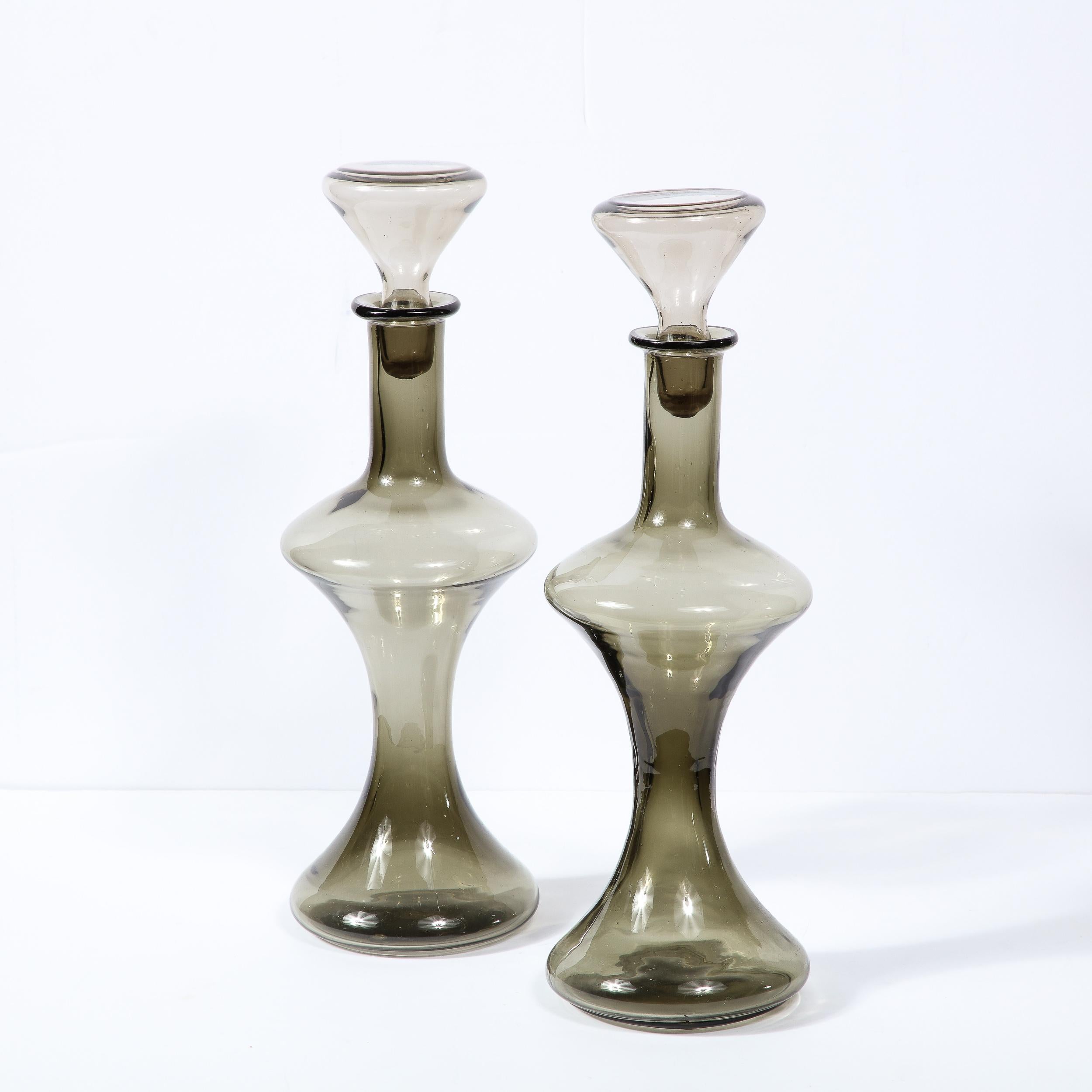 This stunning pair of Mid-Century Modern decanters were realized in Murano, Italy- the island off the coast of Venice renowned for centuries for its superlative glass production. They feature sinuously curved and undulating bodies in a sultry