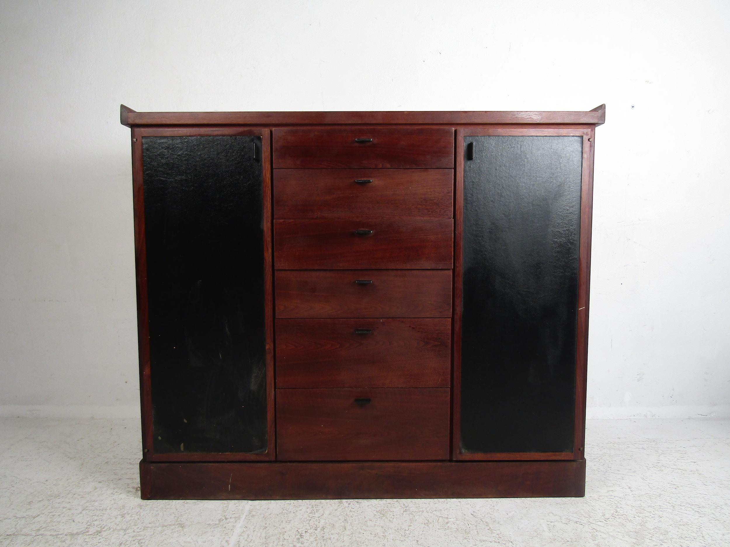 Stunning Mid-Century Modern highboy dresser. Six spacious dovetail-jointed drawers line the center of the case and are flanked by leather front cabinet doors with adjustable shelving within. Raised edges at the case's peak provide a nice accent to