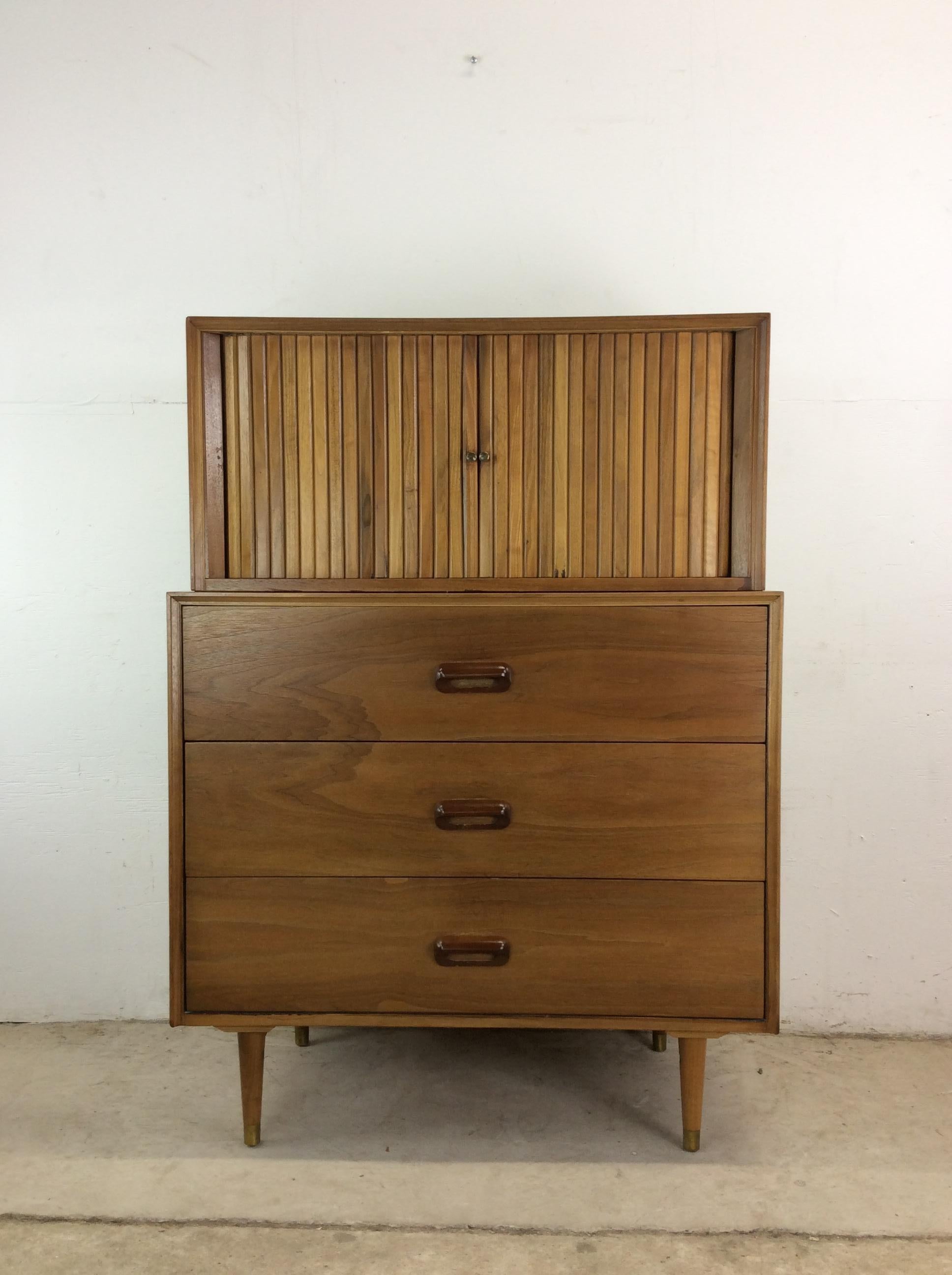 This mid century modern highboy dresser features hardwood construction, walnut veneer with original finish, two piece design with tambour door containing two dovetailed drawers up top and three dovetailed drawers with carved wood hardware, and brass