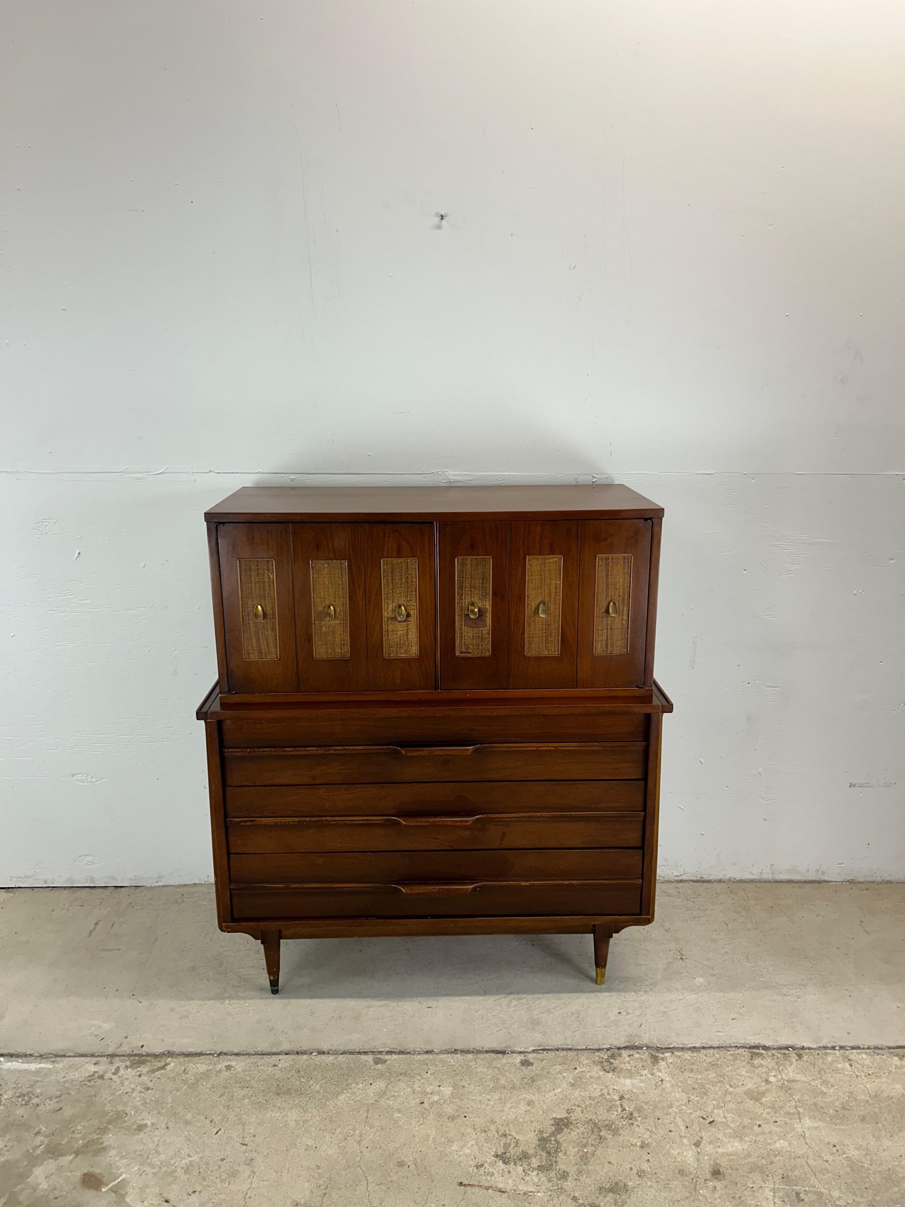 Questo comò highboy della metà del secolo scorso presenta una struttura in legno massiccio, una bella impiallacciatura in noce con finitura originale, cinque cassetti a coda di rondine, due ante del mobile con frontale a canne e ferramenta in
