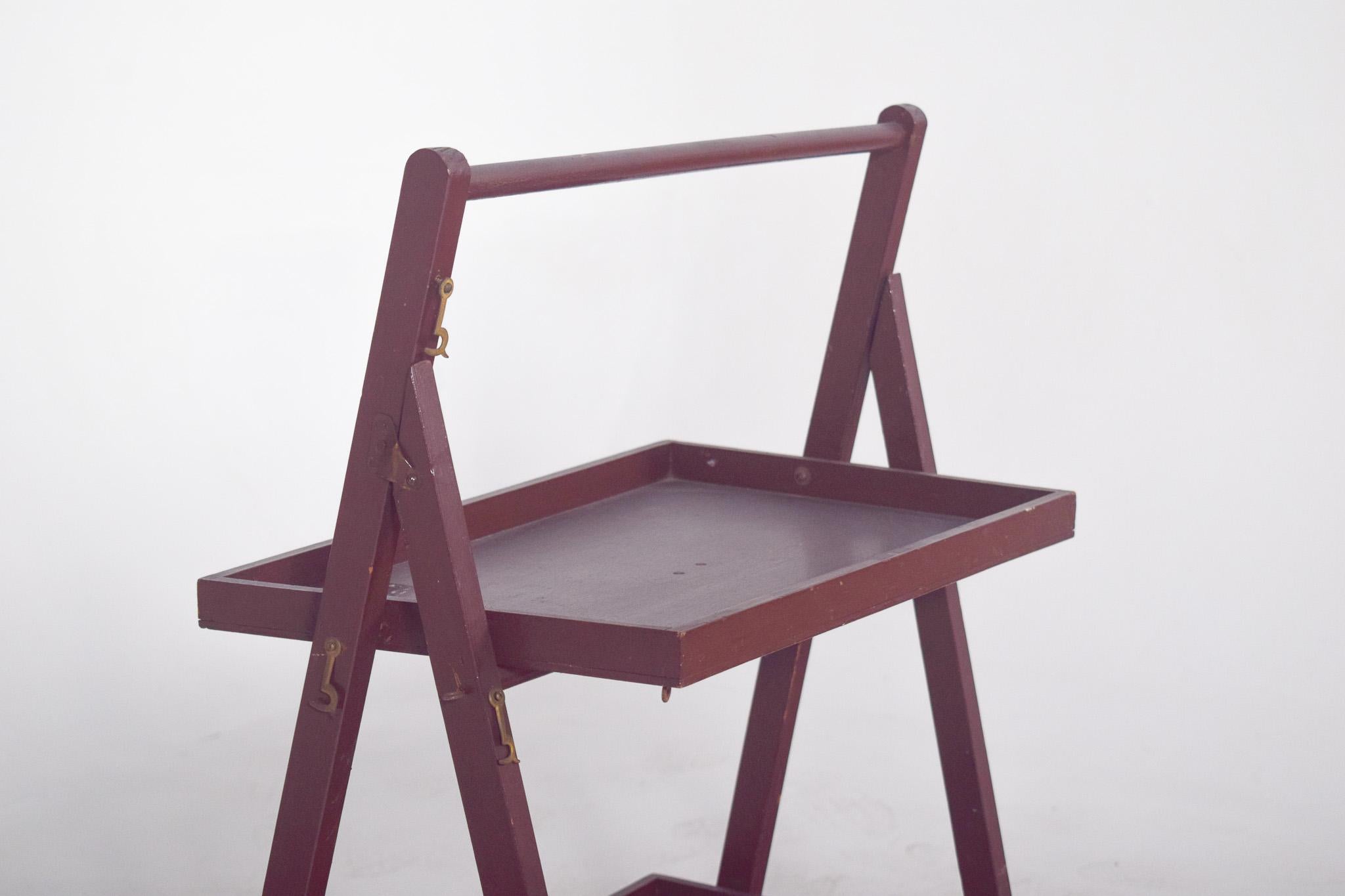 Revistero de madera abatible. Con un diseño funcional y una estructura de caballete plegable, la pieza tiene dos soportes de bandeja, uno superior y otro inferior, ambos sujetos a un bastidor inclinado con juntas metálicas laterales que permiten