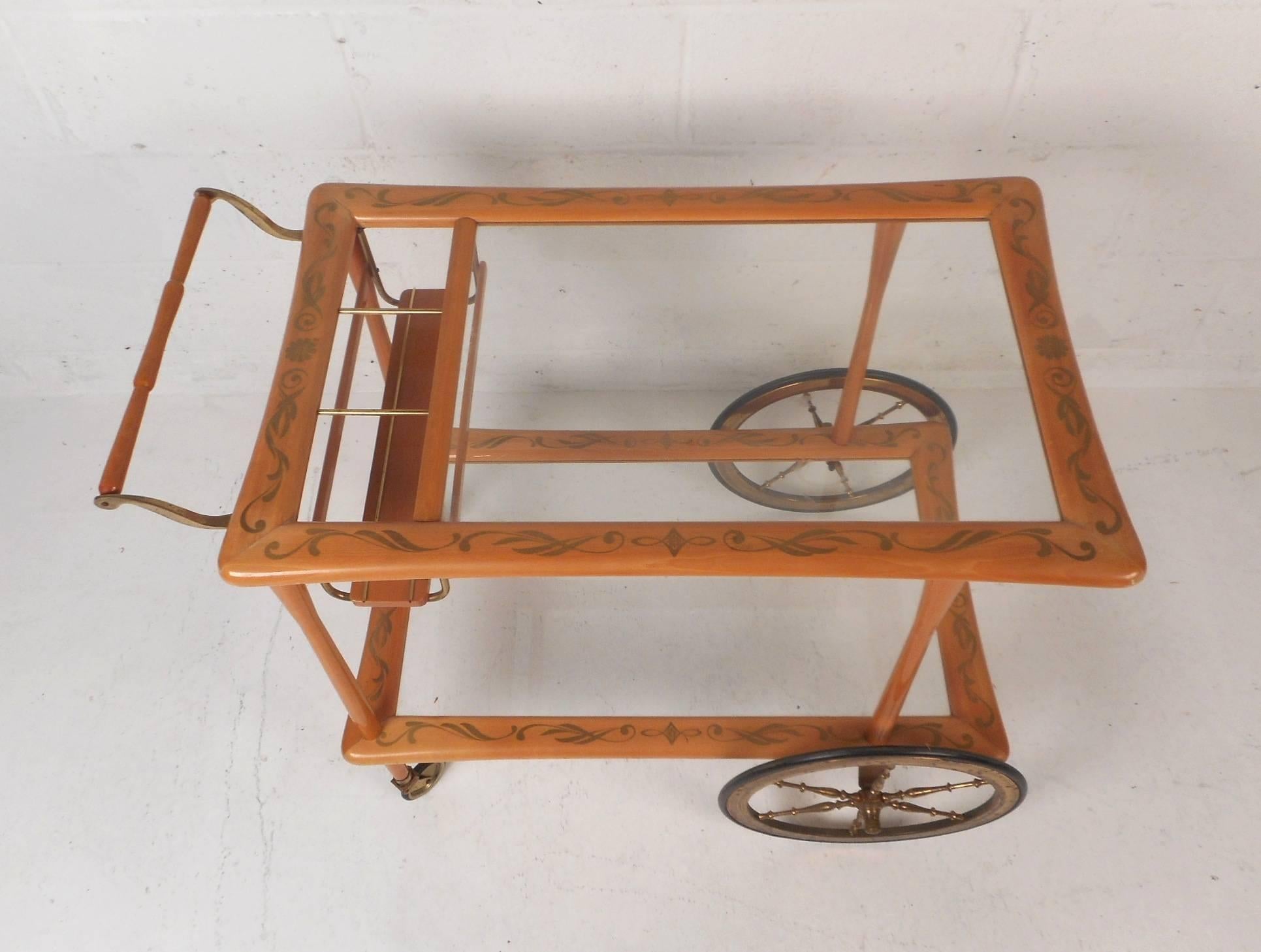 This stunning vintage modern bar cart features two tiers with glass shelves and an unusual bottle holder by the handle. A sturdy frame with fabulous inlaid designs throughout. This unique cart has two oversized front wheels with spoked brass and two