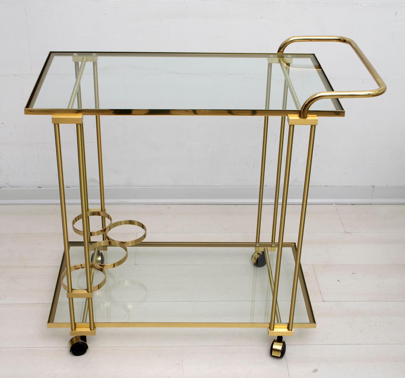 Midcentury brass bar cart, circa 1970s. It is versatile in size and can be used as a bar or serving cart or as an additional storage space in an office. It has four bottle holders at the bottom and two large glass shelves.
Italian production of