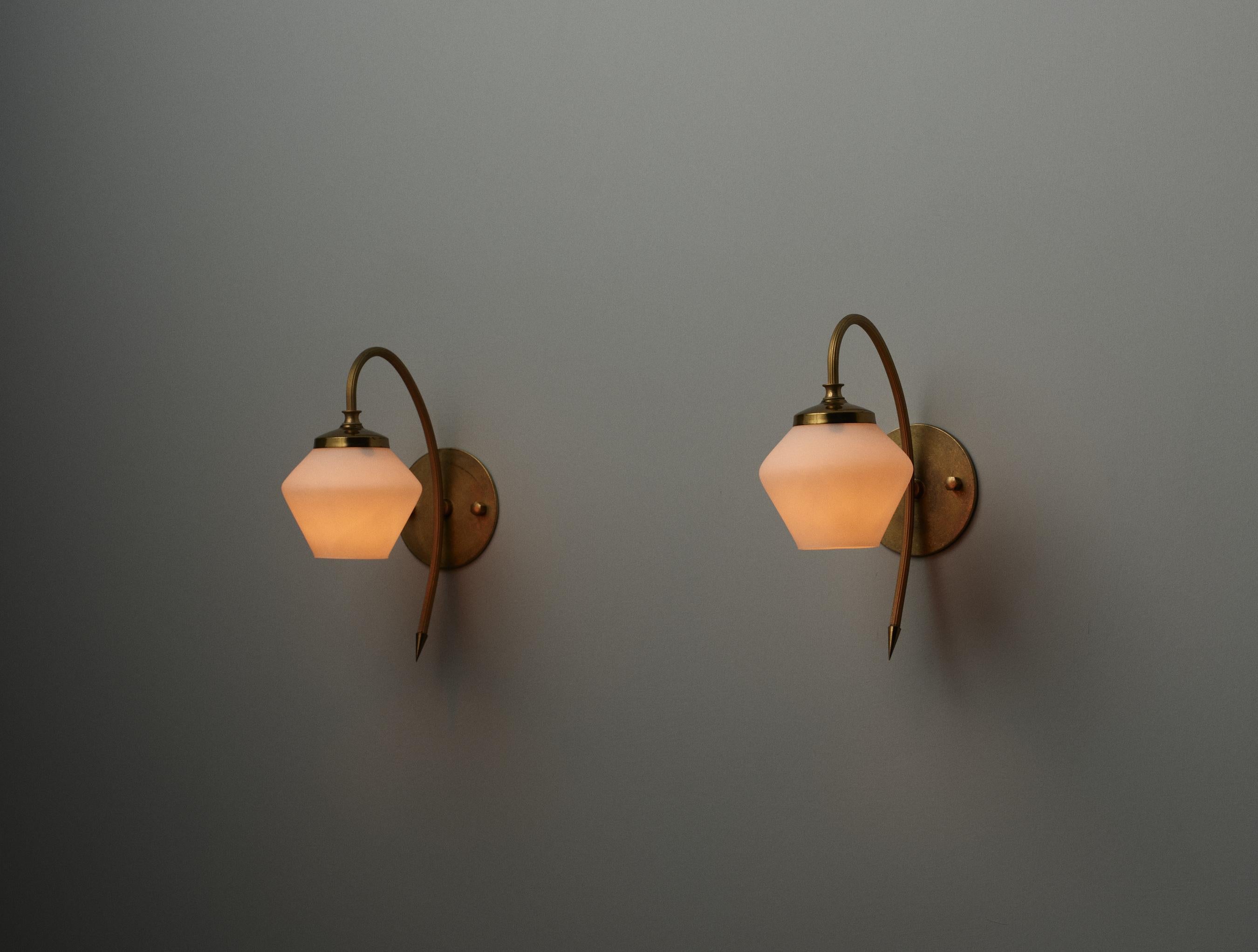 This pair of wall sconces exemplifies Italian design from the 1950s, featuring a clean and elegant aesthetic. Each sconce is crafted from brass, showcasing a well-maintained patina that adds character to the metal's surface. The sconces are equipped