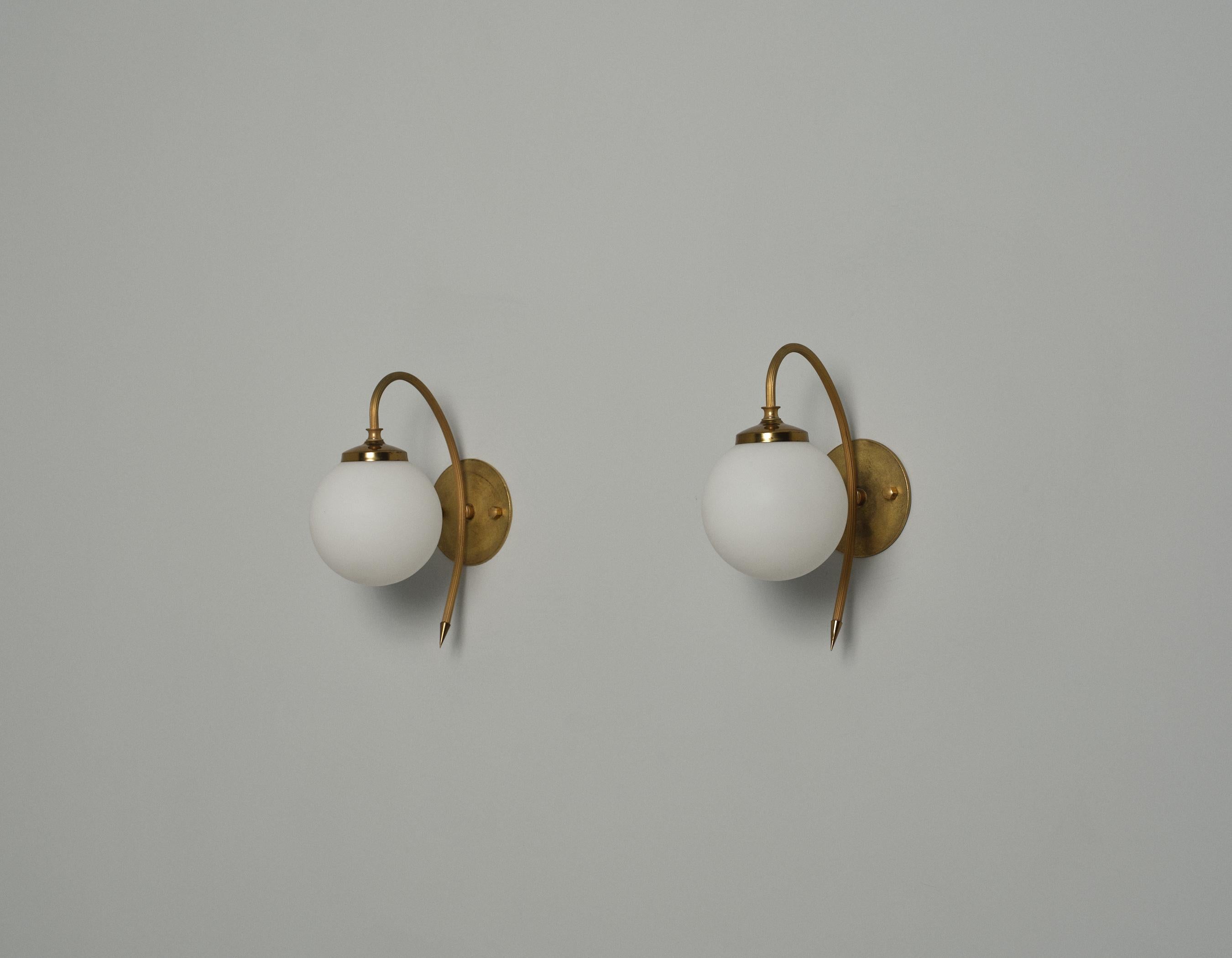 This pair of wall sconces exemplifies Italian design from the 1950s, featuring a clean and elegant aesthetic. Each sconce is crafted from brass, showcasing a well-maintained patina that adds character to the metal's surface. The sconces are equipped