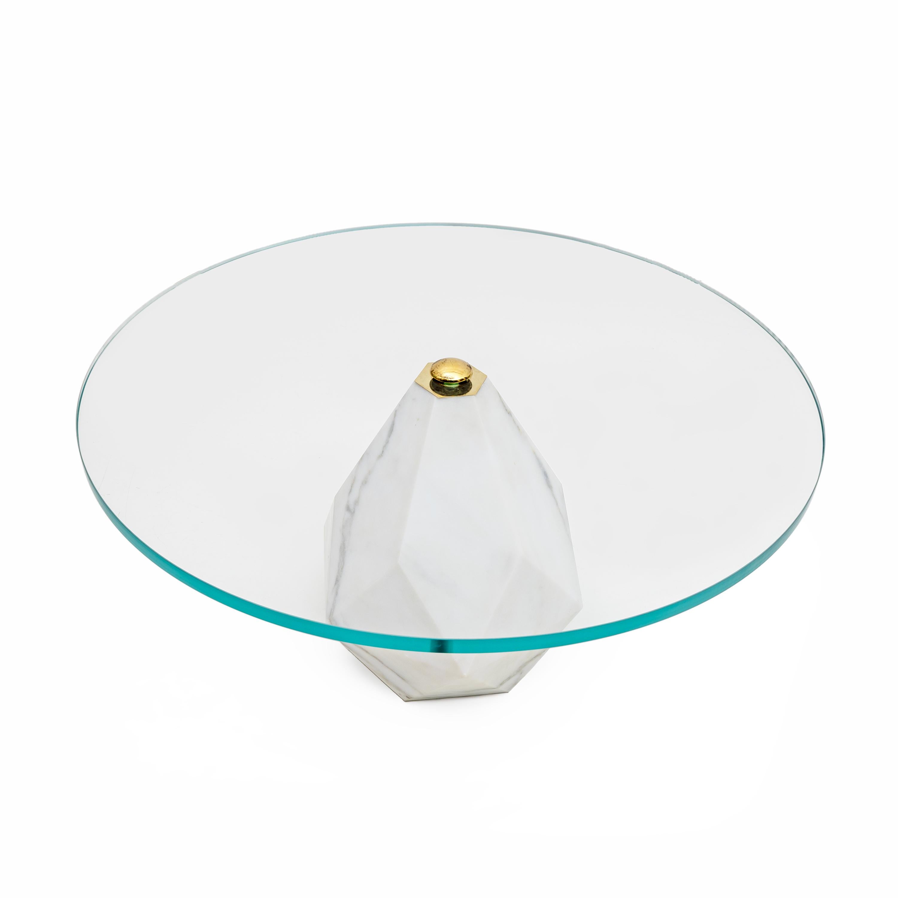 Modern minimalist coffee table with a thick circular top made of clear glass. The central base of the table is made of grey veined white marble, Carrara marble, with a geometric and faceted design that adds visual interest and elegance. A screw