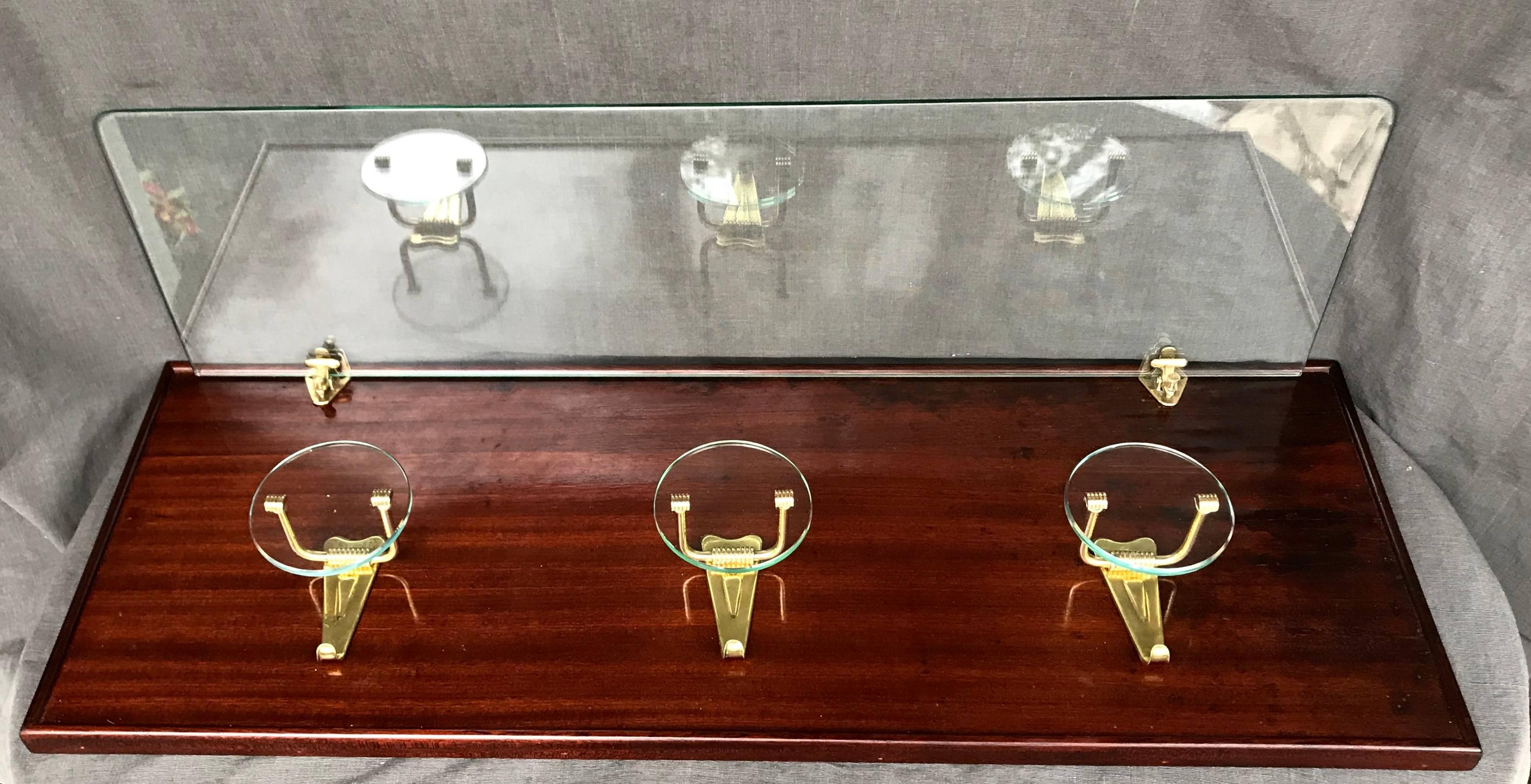 Mid-Century Modern Italian coat and hat rack with shelf.  Mahogany panel of spare design supporting three glass and brass coat hooks beneath brass clips supporting a glass shelf for hats. Italy, circa 1940.
Dimensions: 34