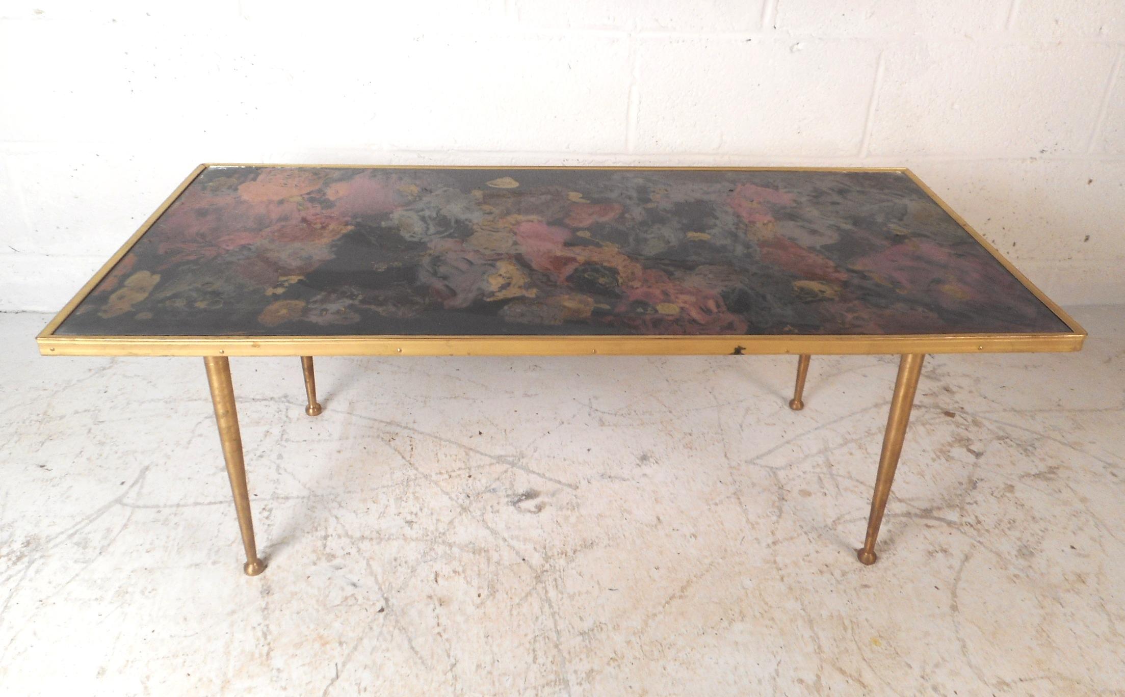 This stunning vintage modern coffee table features a colorful abstract painting on the top underneath a rectangular piece of glass. A sleek Italian design with brass trim along the sides and tapered brass legs with ball feet. An articulate piece