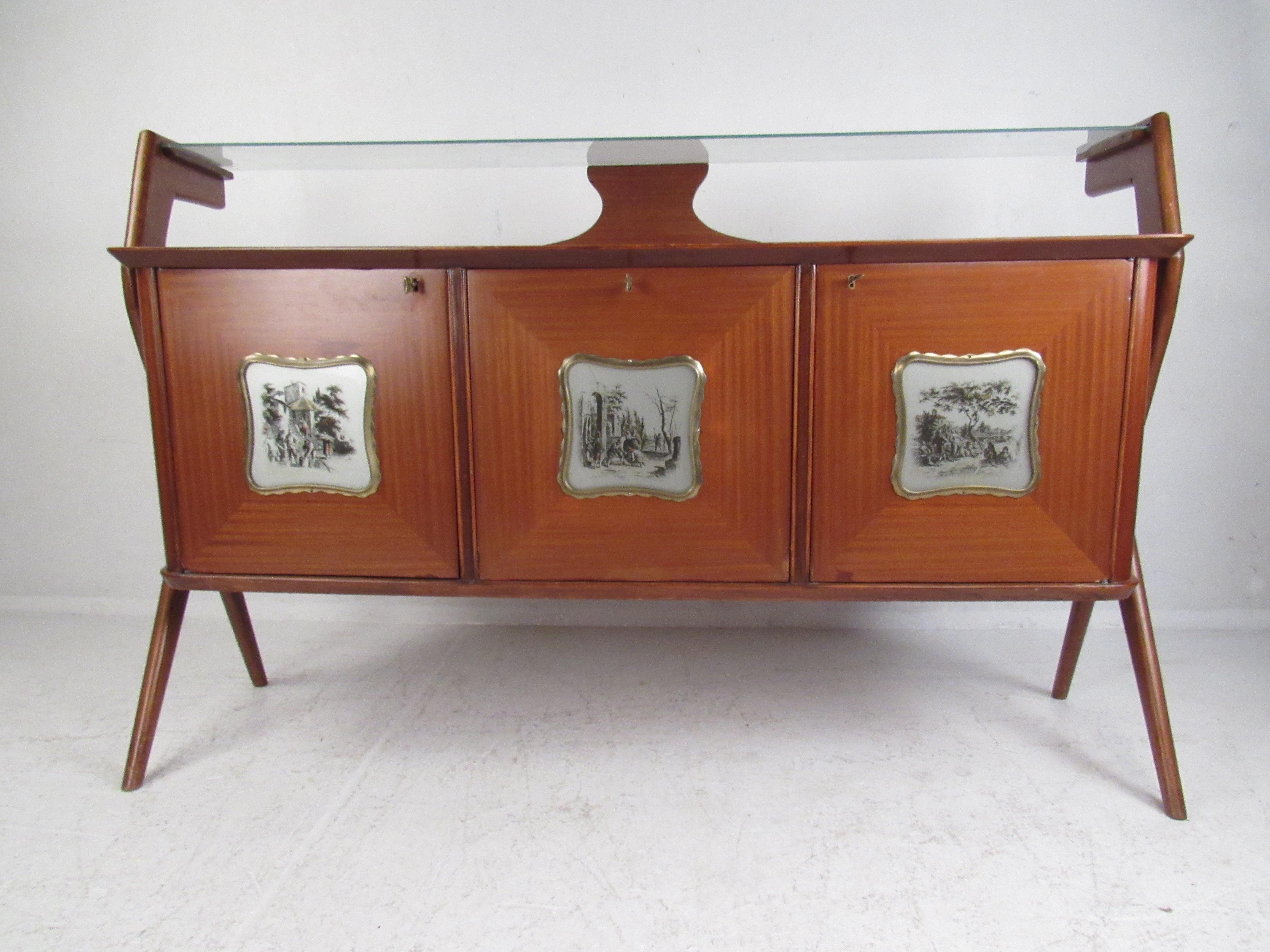 This stunning vintage modern sideboard features a floating glass top and angled legs. Sleek design with three decorative cabinet doors that depict old fashioned European scenery painted on glass fixtures. Lovely vintage finish with elegant wood