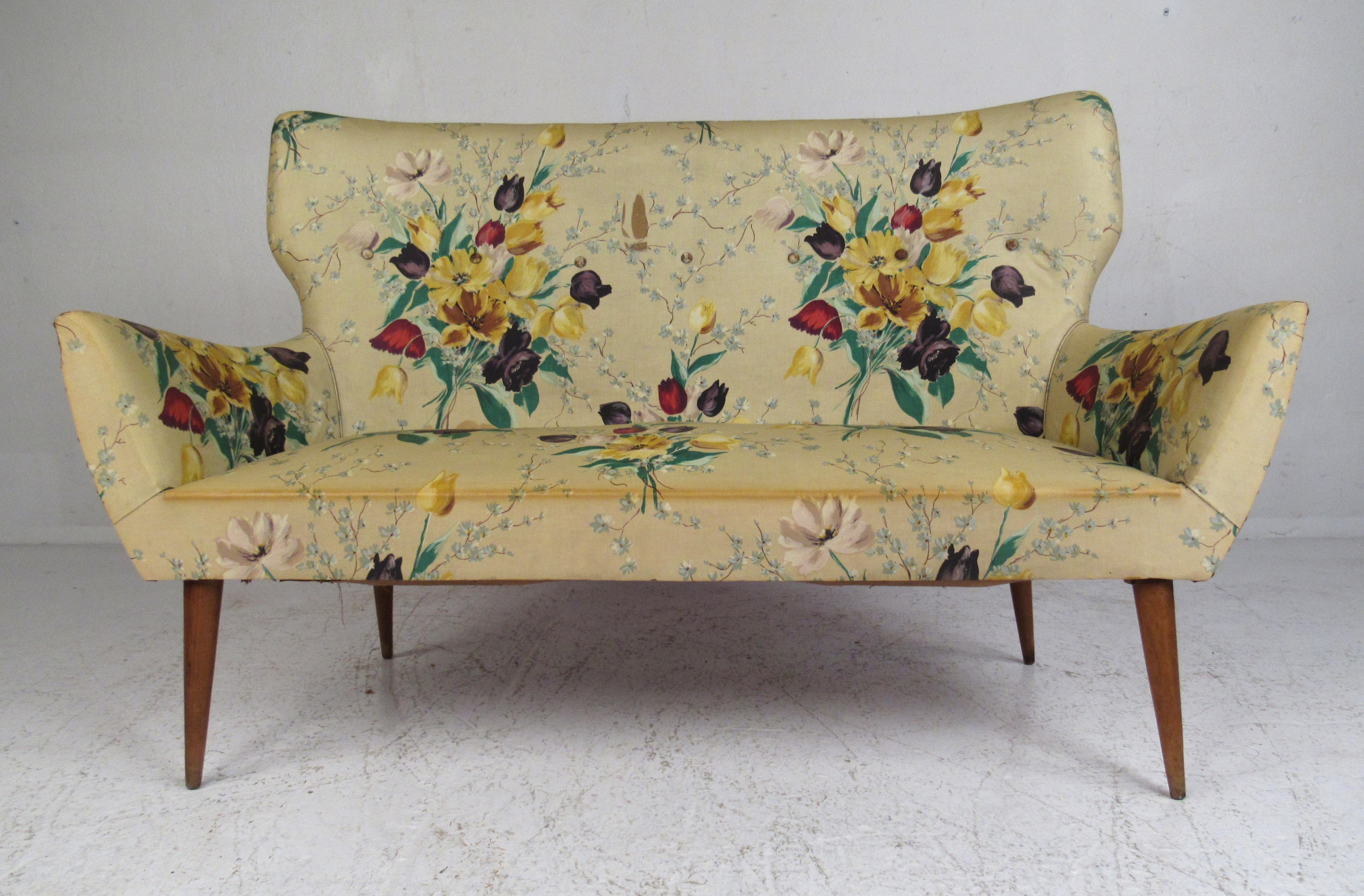 This stunning vintage modern settee features tapered walnut legs and decorative floral upholstery. The unique winged backrest and armrests are sure to make this piece stand out in any seating arrangement. Please confirm item location (NY or