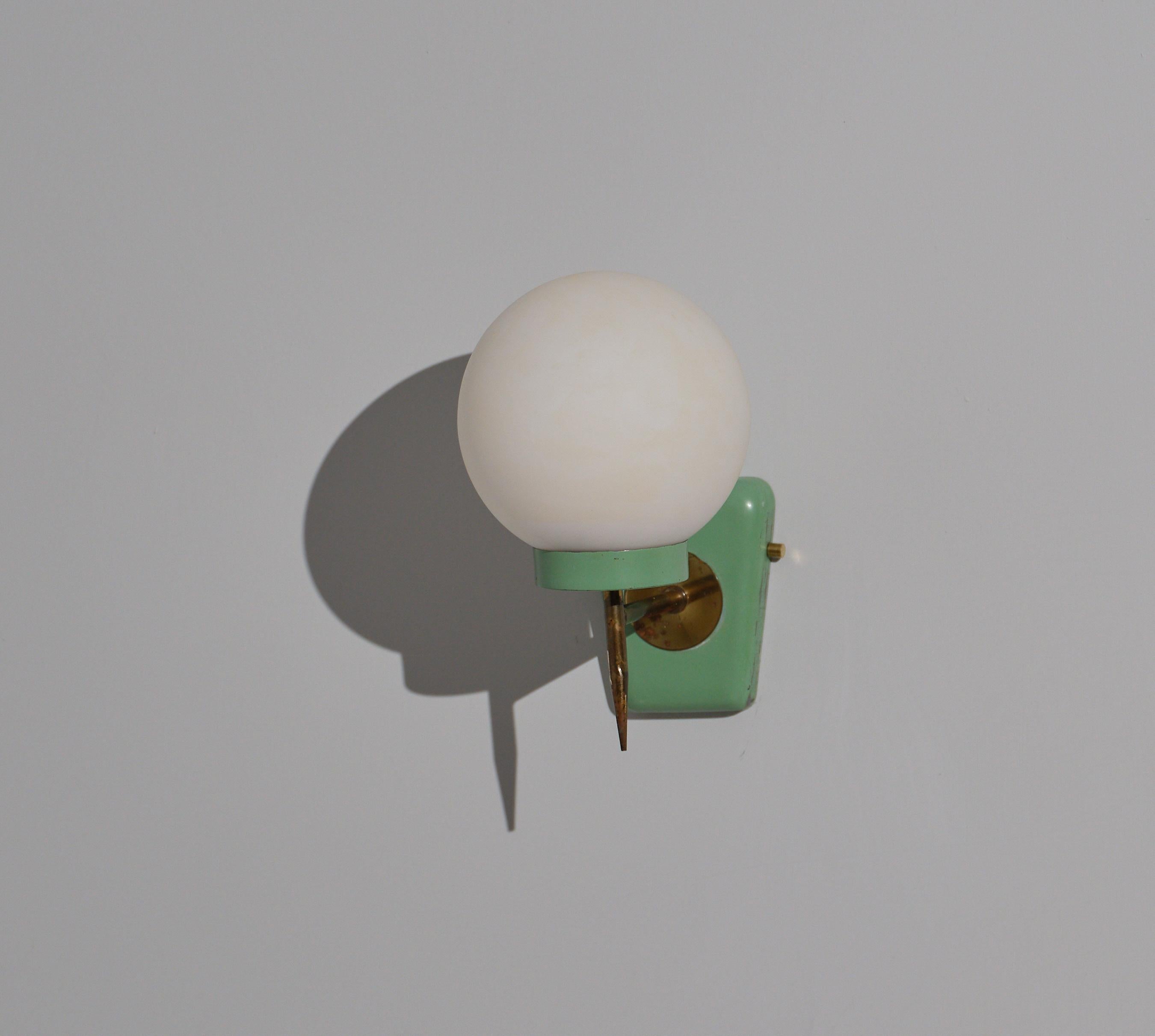 This wall-mounted sconce exemplifies Italian design from the 1950s, featuring a minimalist yet functional aesthetic. The fixture is composed of a single opaline glass sphere that diffuses light softly, mounted on a bracket made from enameled steel