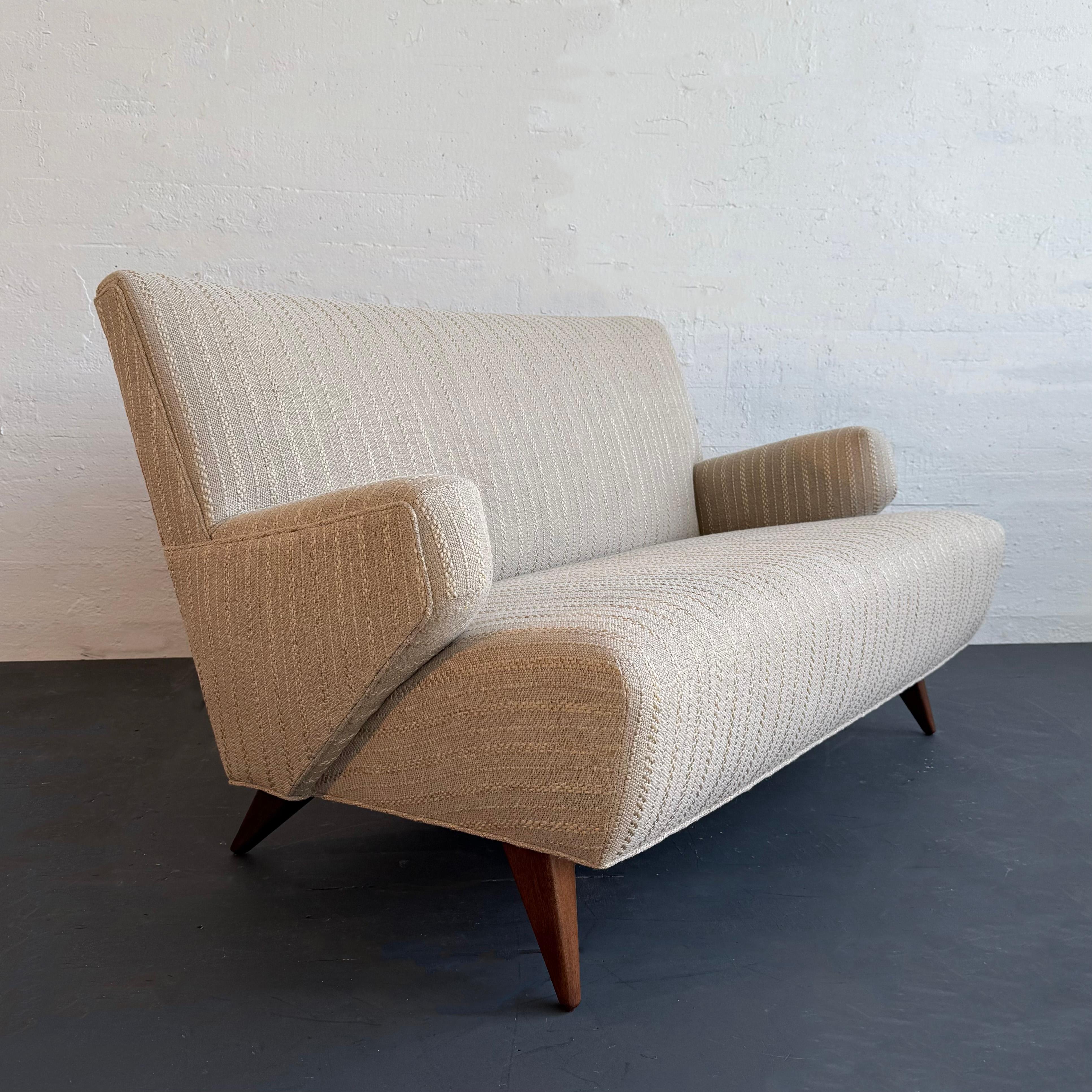 This early Jens Risom model 37 loveseat sofa for Knoll is a classic example of Mid-Century Modern design. The fully upholstered sofa features a high back with rounded edges and recessed arms that follow the angle of the tapered splayed stained birch