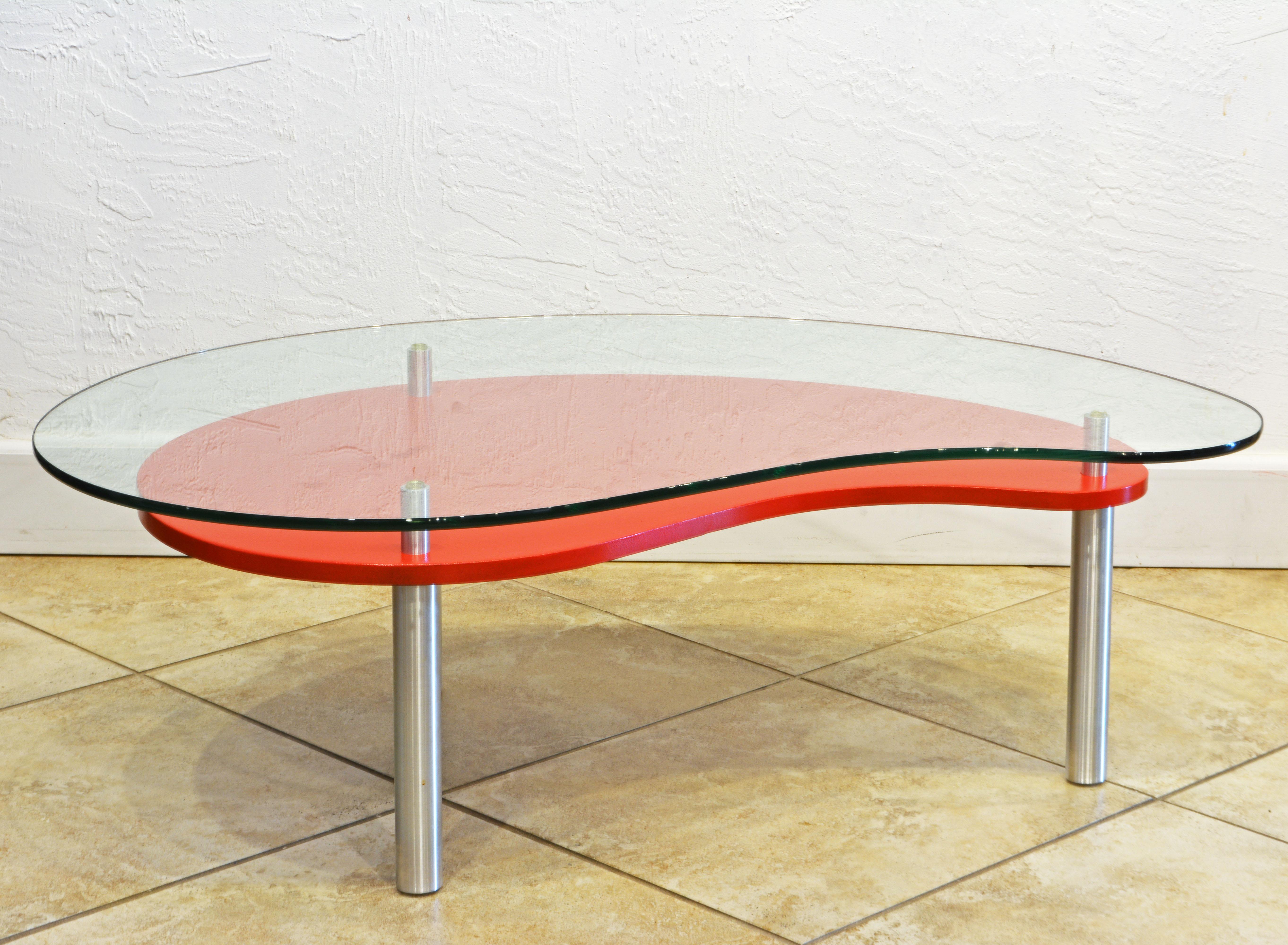 This Minimalist design kidney shape two tier coffee table features a thick tempered glass top on brushed aluminum legs and a lower red painted display shelf adding an almost abstract effect.