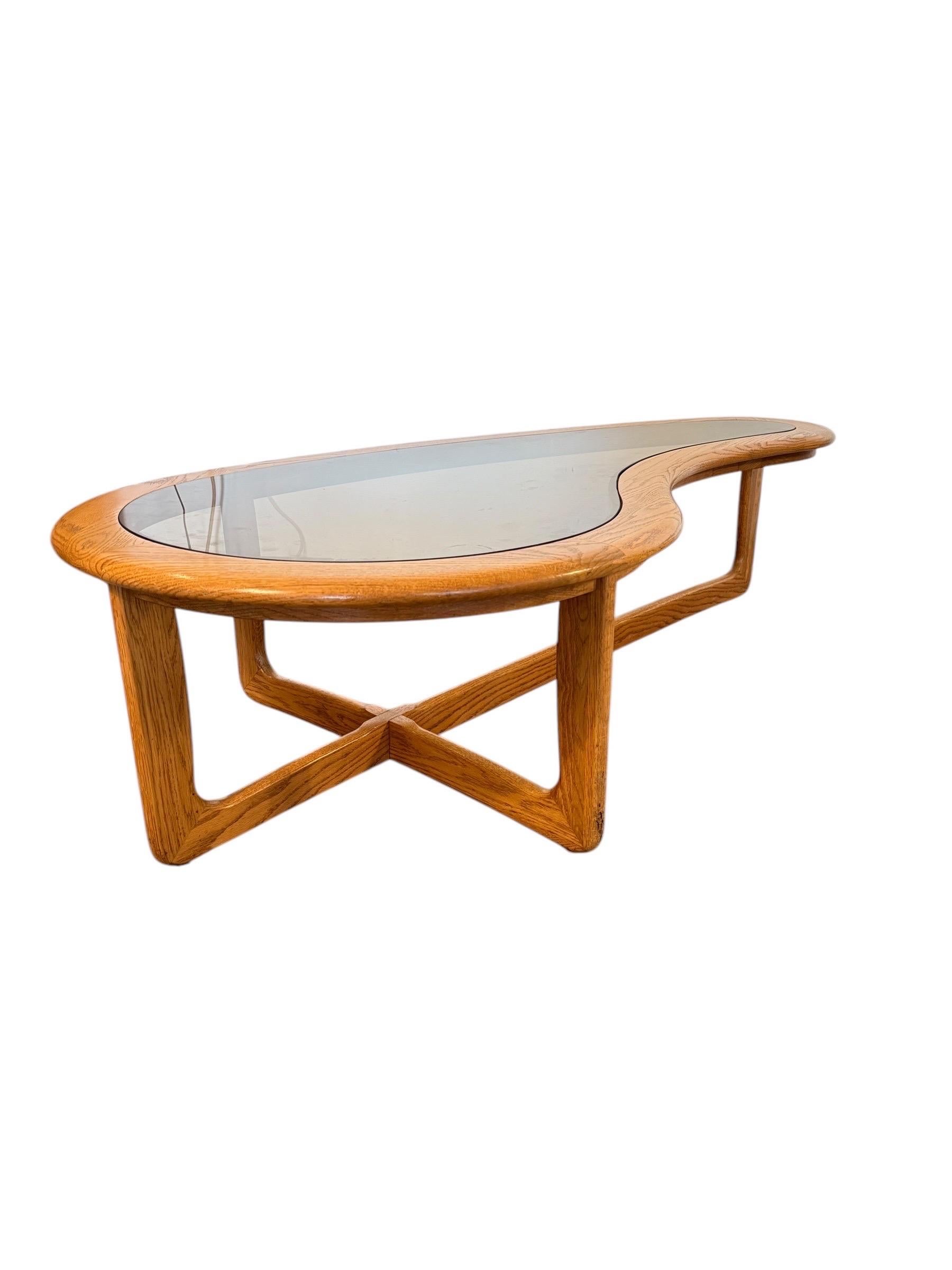 Llamativa desde todos los ángulos, esta escultural mesa de centro moderna de mediados de siglo de Lane Furniture presenta una icónica silueta en forma de riñón combinada con una tapa de cristal ahumado. La base de roble en forma de X proporciona