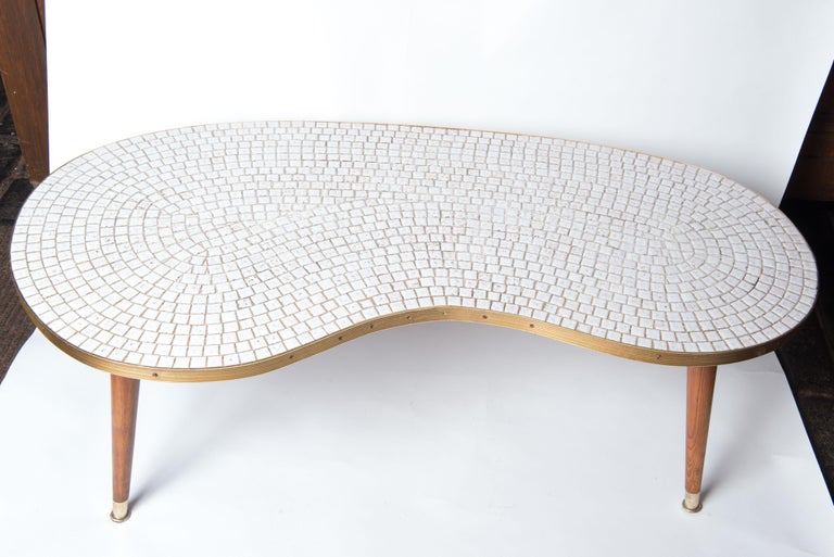 Mid-Century Modern kidney shaped glazed white ceramic tile top coffee table, tapered wood legs with brass caps. The curved perimeter of the table top is edged with a brass band. The glazed white ceramic tile top is in excellent vintage condition.
