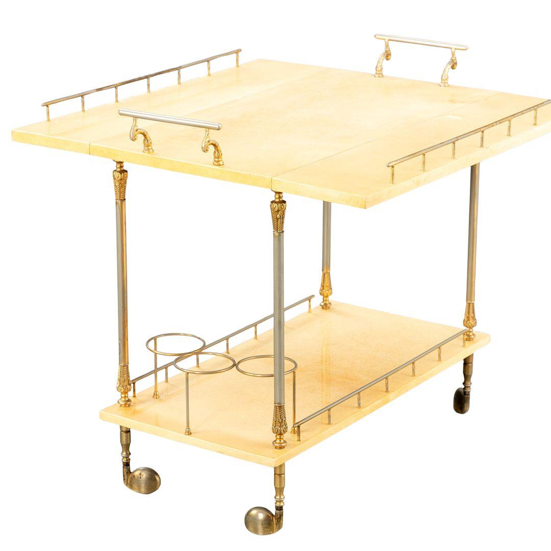 Aldo Tura designed this bar cart/serving trolley, featuring a brass frame and two wooden shelves covered in beige-colored goatskin parchment, delicately painted. Its dimensions measure approximately Height 74 cm x Width 80 cm x Depth 42.5/74 cm.