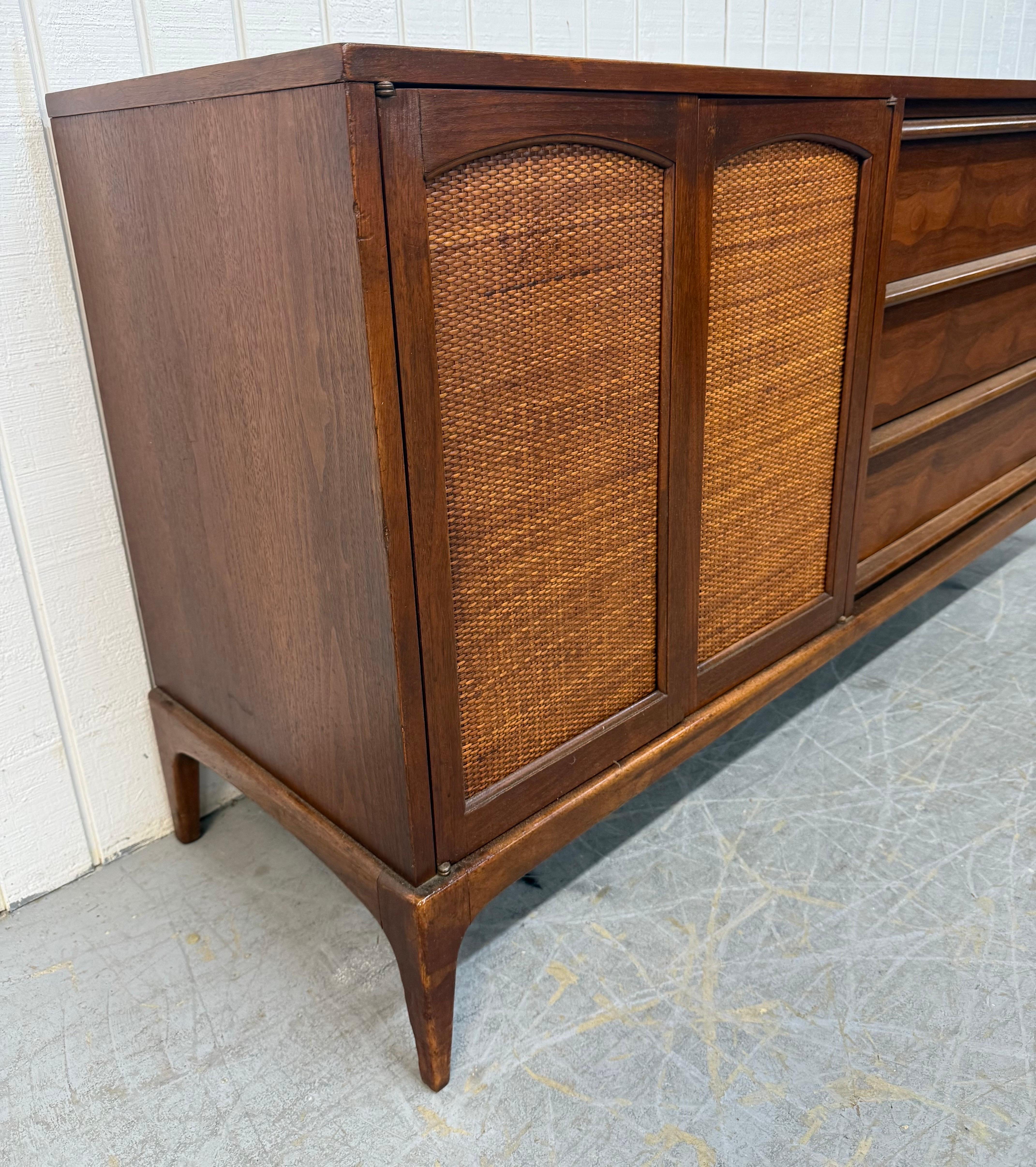 Cette annonce concerne un buffet en noyer Lane Rhythm Mid-Century Modern. Ce meuble présente un design rectiligne, deux portes en rotin qui s'ouvrent sur un tiroir caché avec un espace de rangement, trois tiroirs avec des poignées en bois sur le