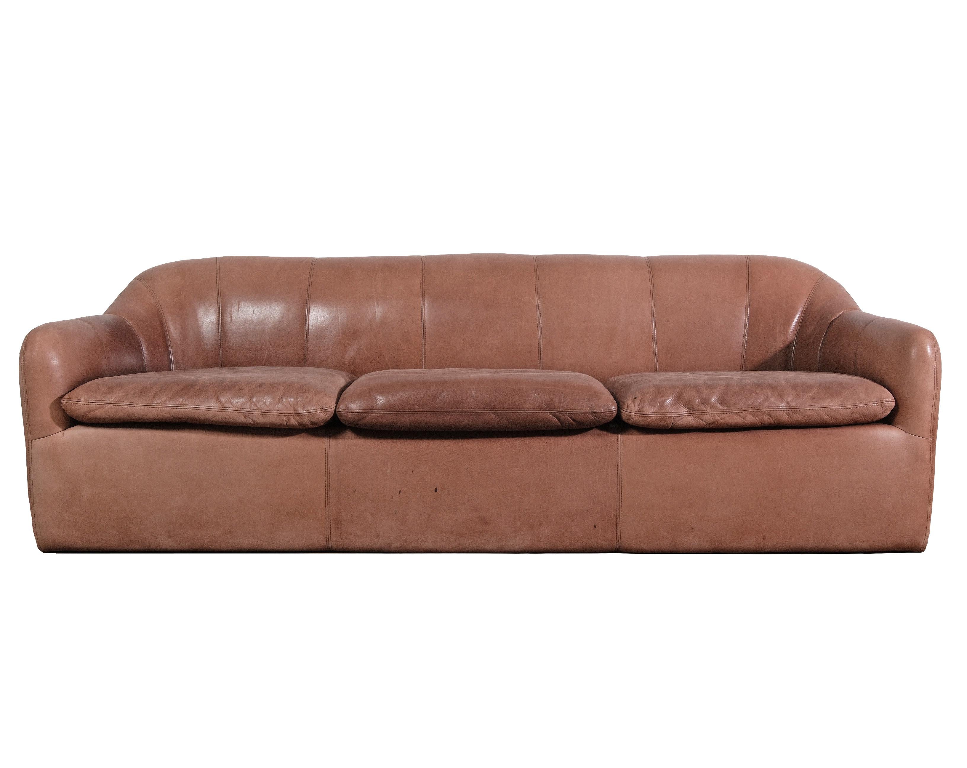 Questo divano in pelle marrone, splendidamente rodato, incarna elementi di design moderno della metà del secolo scorso e porta il tuo spazio negli anni '70. Caratterizzato da un lussuoso rivestimento in pelle cognac con finitura patinata, il divano