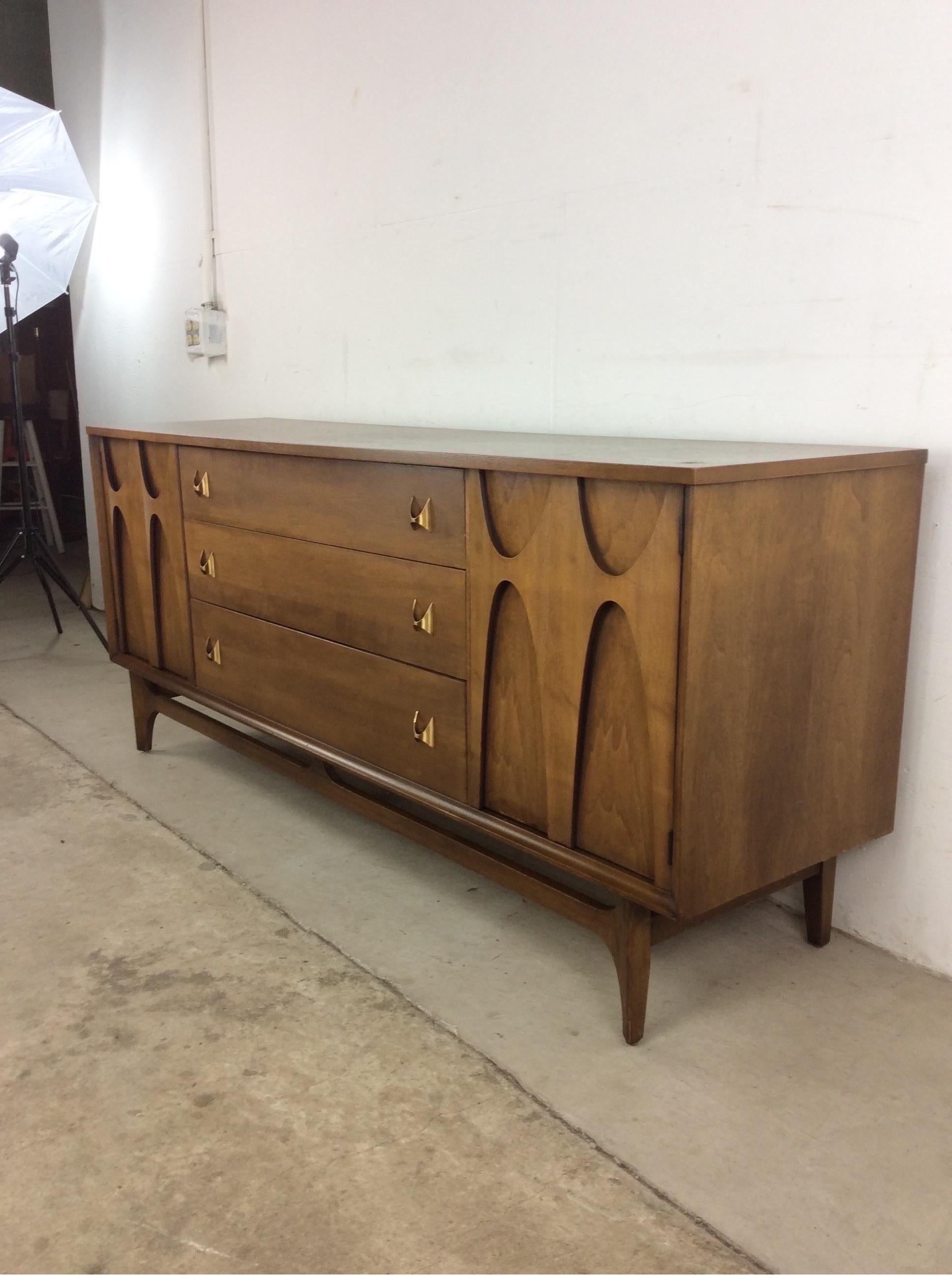 This Mid-Century Modern lowboy dresser from the Brasilia line by Broyhill features hardwood construction, original walnut finish, nine dovetailed drawers, two cabinet doors with signature bentwood detail, brass hardware, and tall tapered legs.