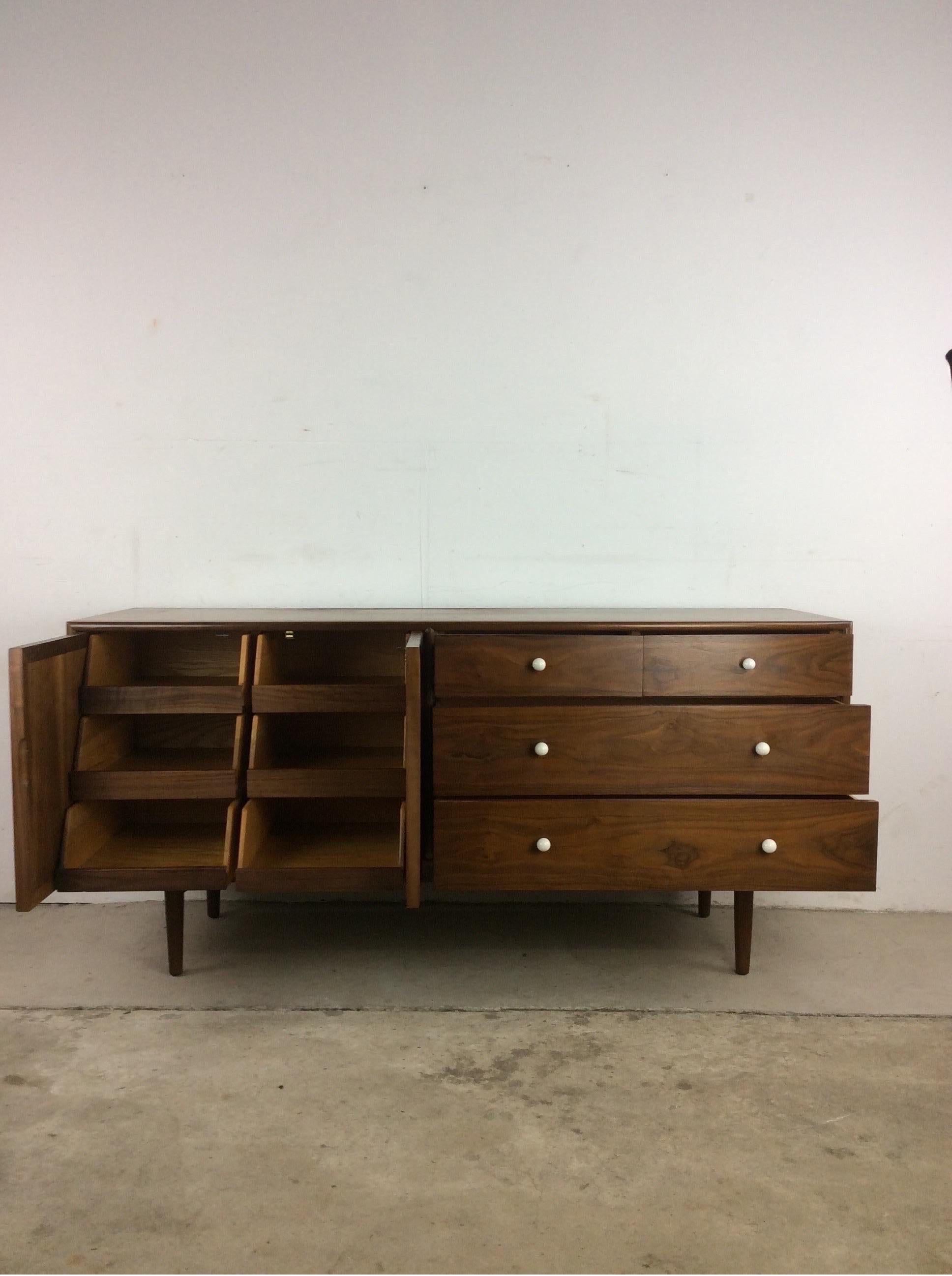 This Mid-Century Modern lowboy dresser from the Declaration line by Drexel features hardwood construction, walnut veneer with original finish, nine dovetailed drawers, two cabinet doors, signature white porcelain hardware, and tall tapered