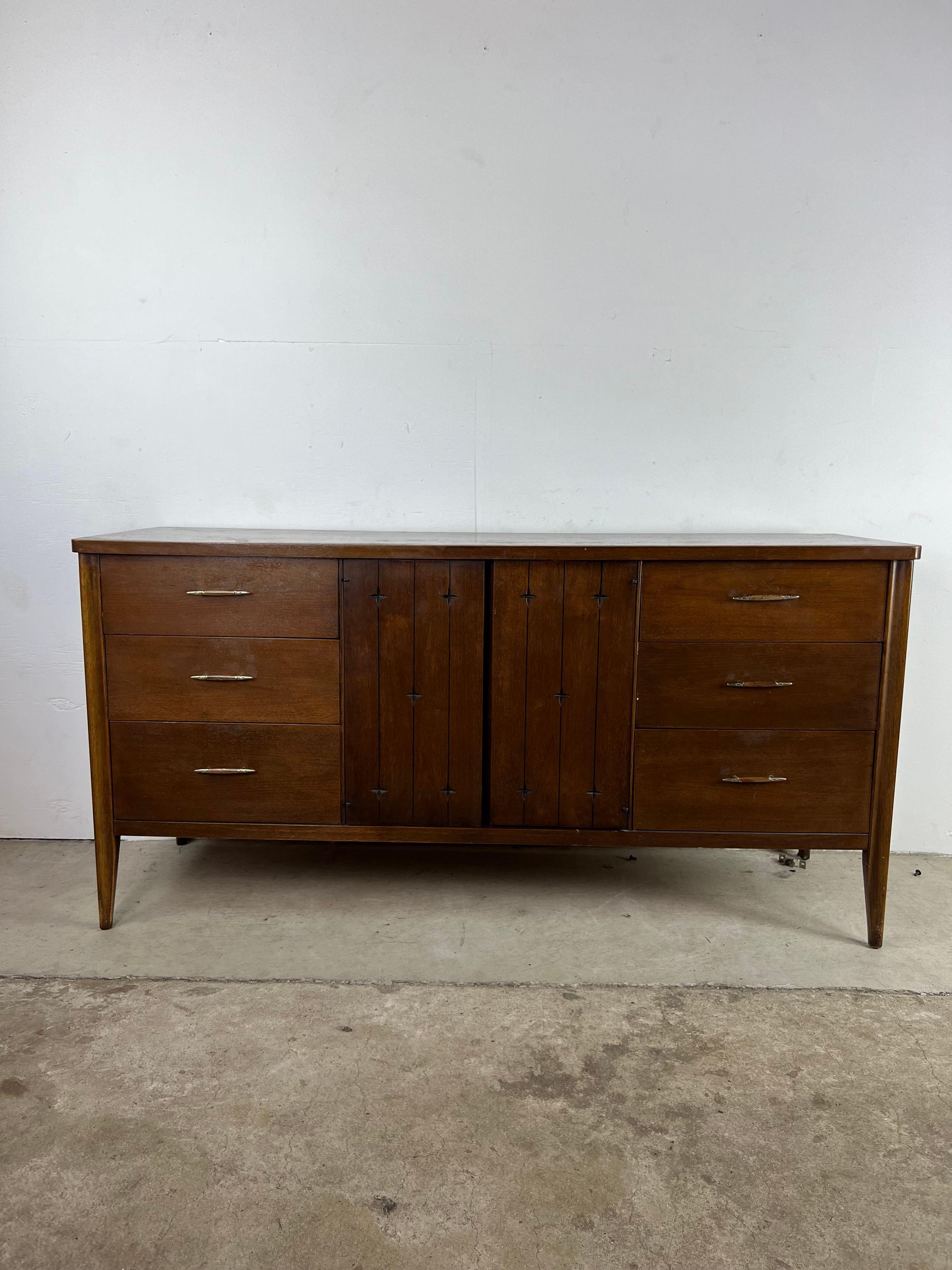This Mid-Century Modern lowboy dresser from the SAGA line by Broyhill features hardwood construction, original walnut finish, nine dovetailed drawers, two cabinet doors with carved cross detailing, and tall tapered legs.

 