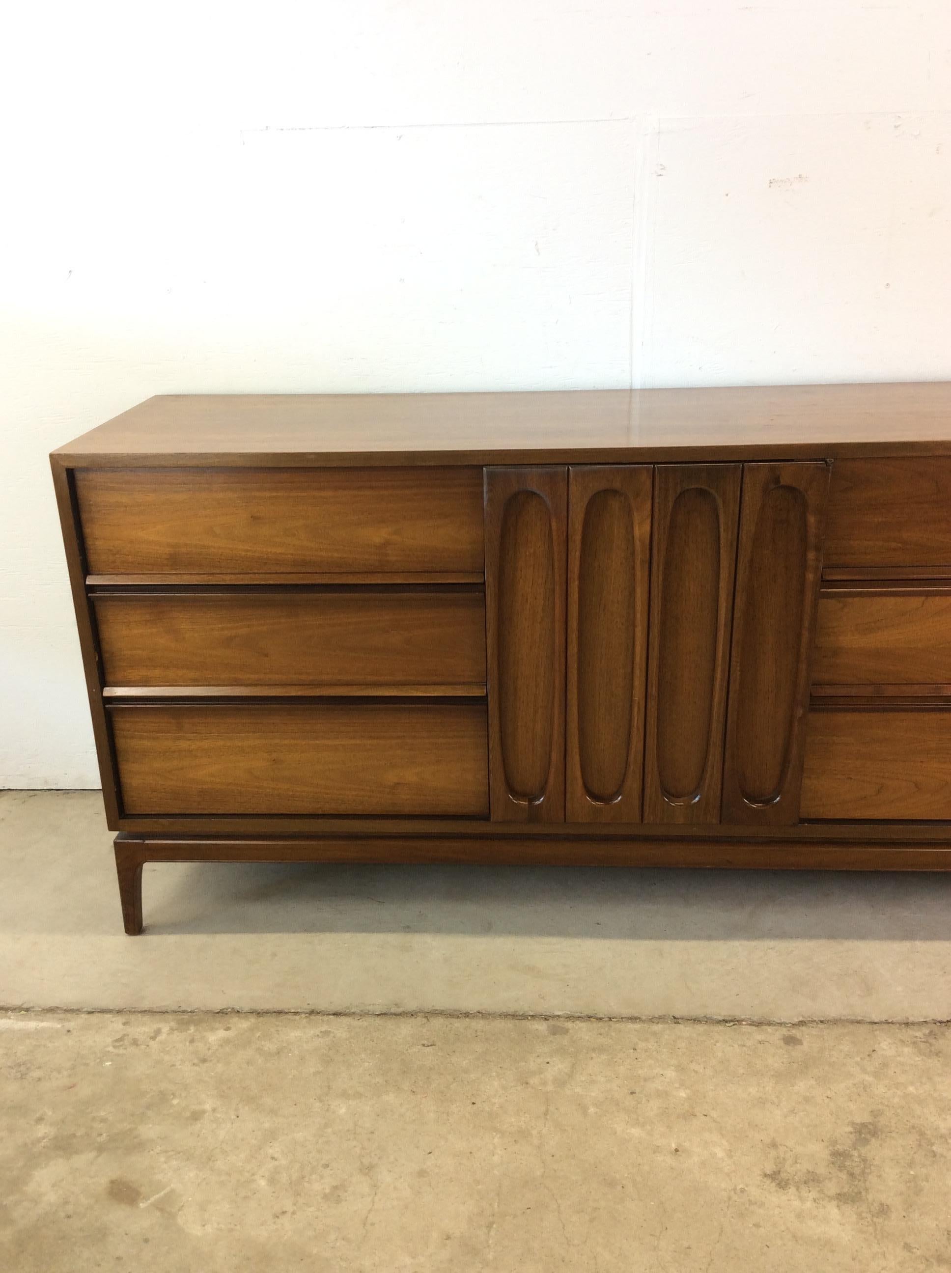 This mid century modern lowboy dresser features hardwood construction, walnut veneer with original finish, nine dovetailed drawers, unique curved wood cabinet doors, and tall tapered legs.  Matching pair of nightstands available separately.