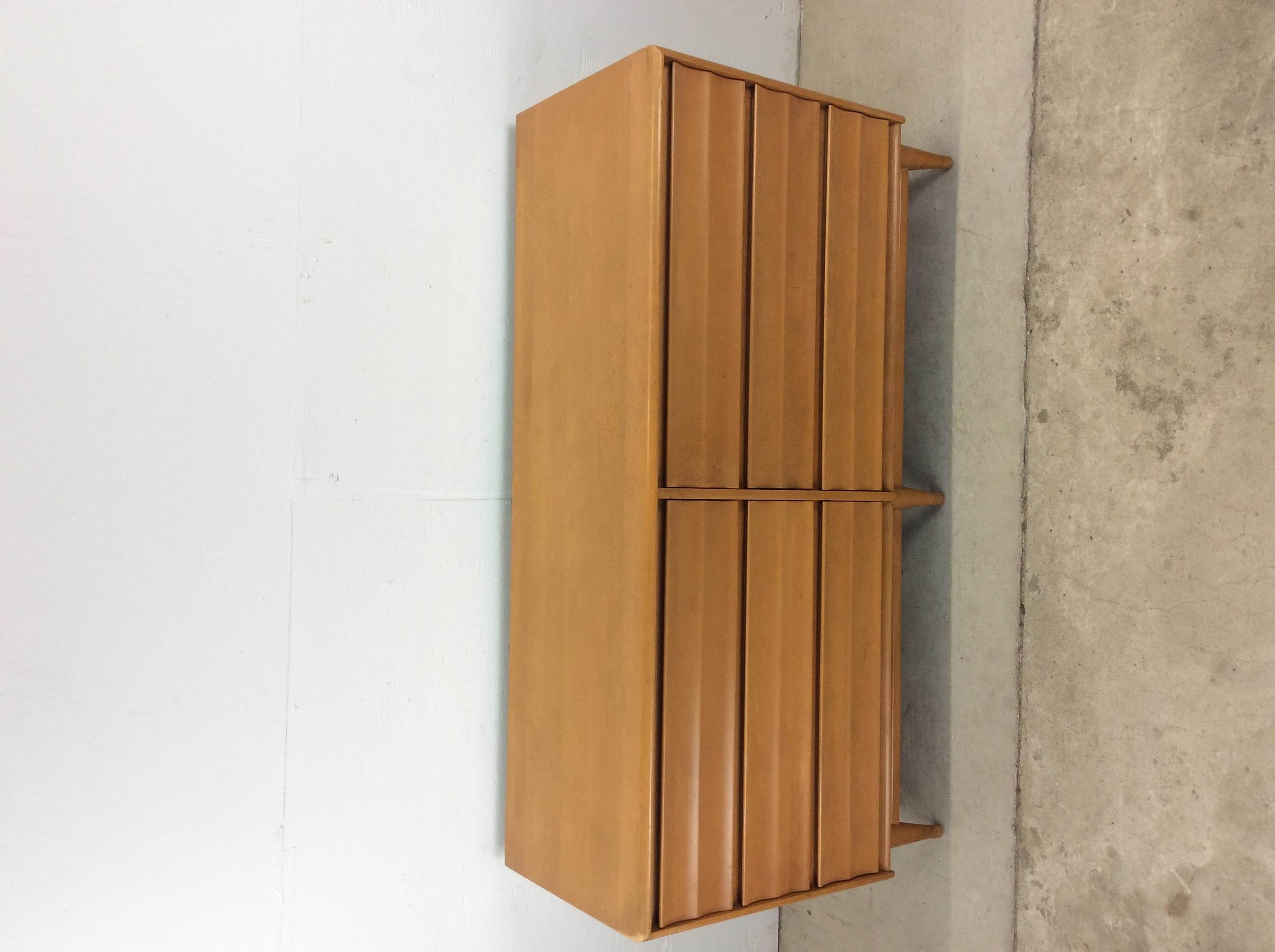 This mid century modern lowboy dresser by Heywood Wakefield features hardwood construction with original champagne finish, six dovetailed drawers with unique louvered front design, mounted mirror with matching finish, and stretcher base with tapered