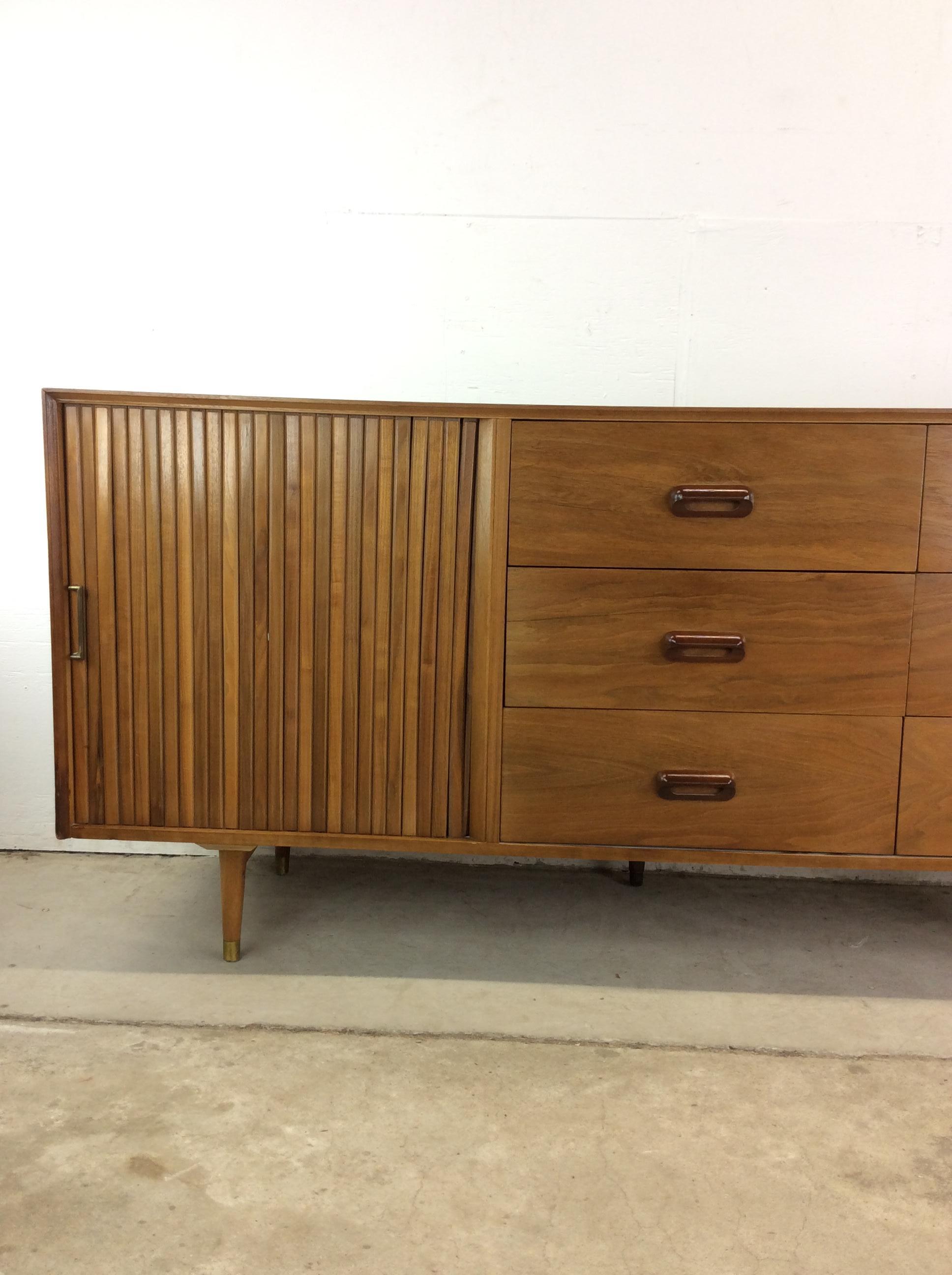 This mid century modern lowboy dresser features hardwood construction, walnut veneer with original finish, tambour door containing three dovetailed drawers, six additional dovetailed drawers with carved wood hardware, and brass capped tapered