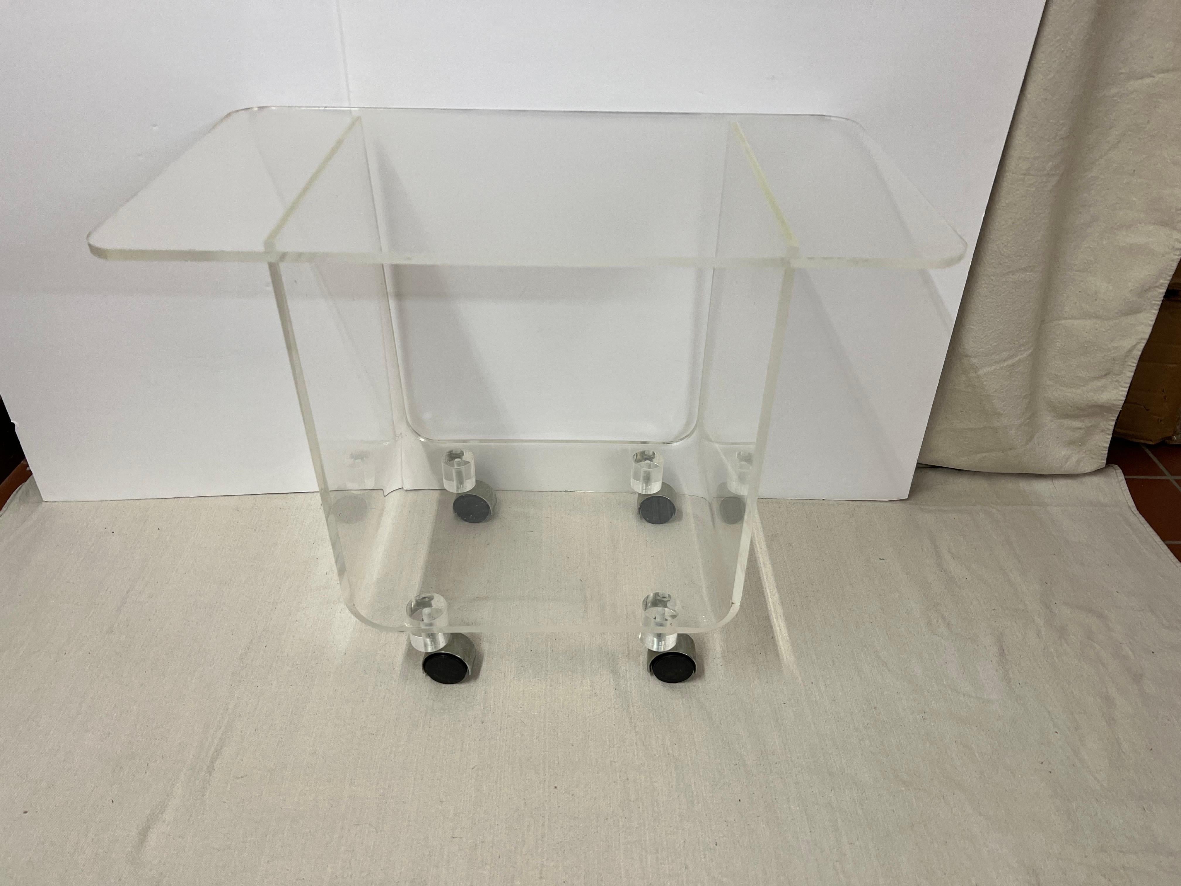 Mid-Century Modern Lucite stand or cart. Post modern vibe to this minimalist cart. Use as a bar cart with bottles on top or underneath or as as a serving stand. Classic black plastic wheels.  Interior shelf measures: 15” wide x 17' tall 
