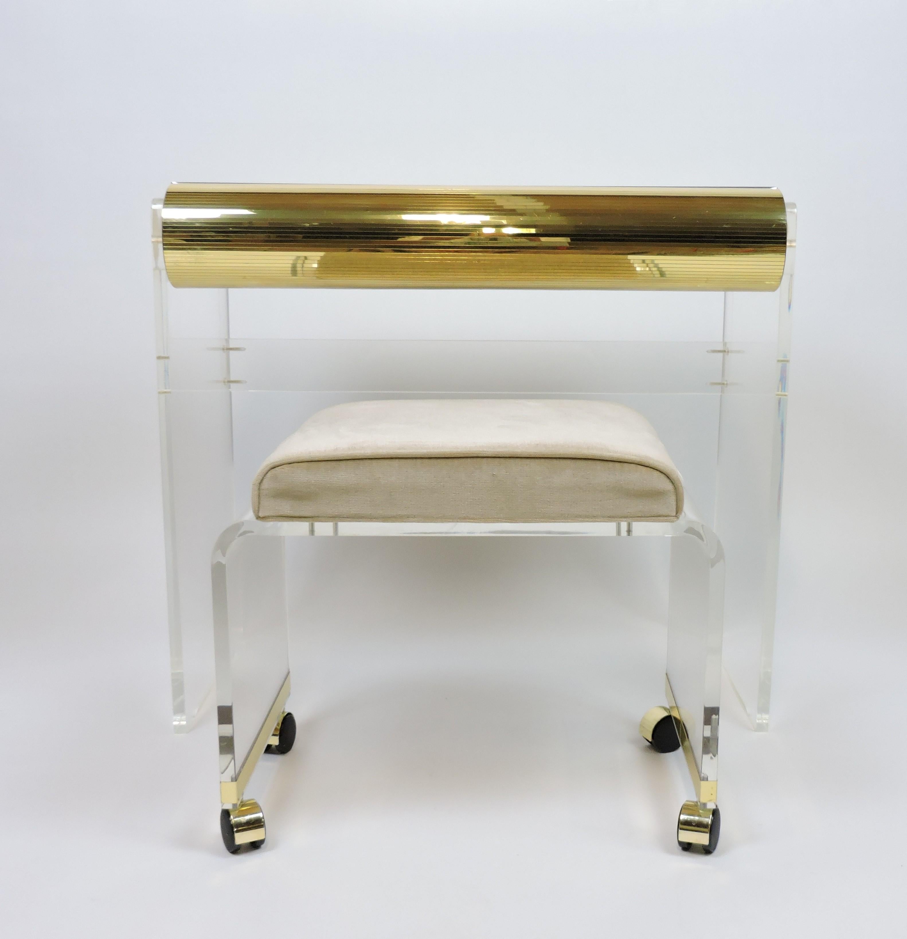 Beautiful Lucite vanity and stool set with brass colored accents. The vanity has thick clear Lucite sides, a mirrored top, and a drawer with a faceted front. The stool is upholstered in a light tan fabric with casters for easy movement and has a