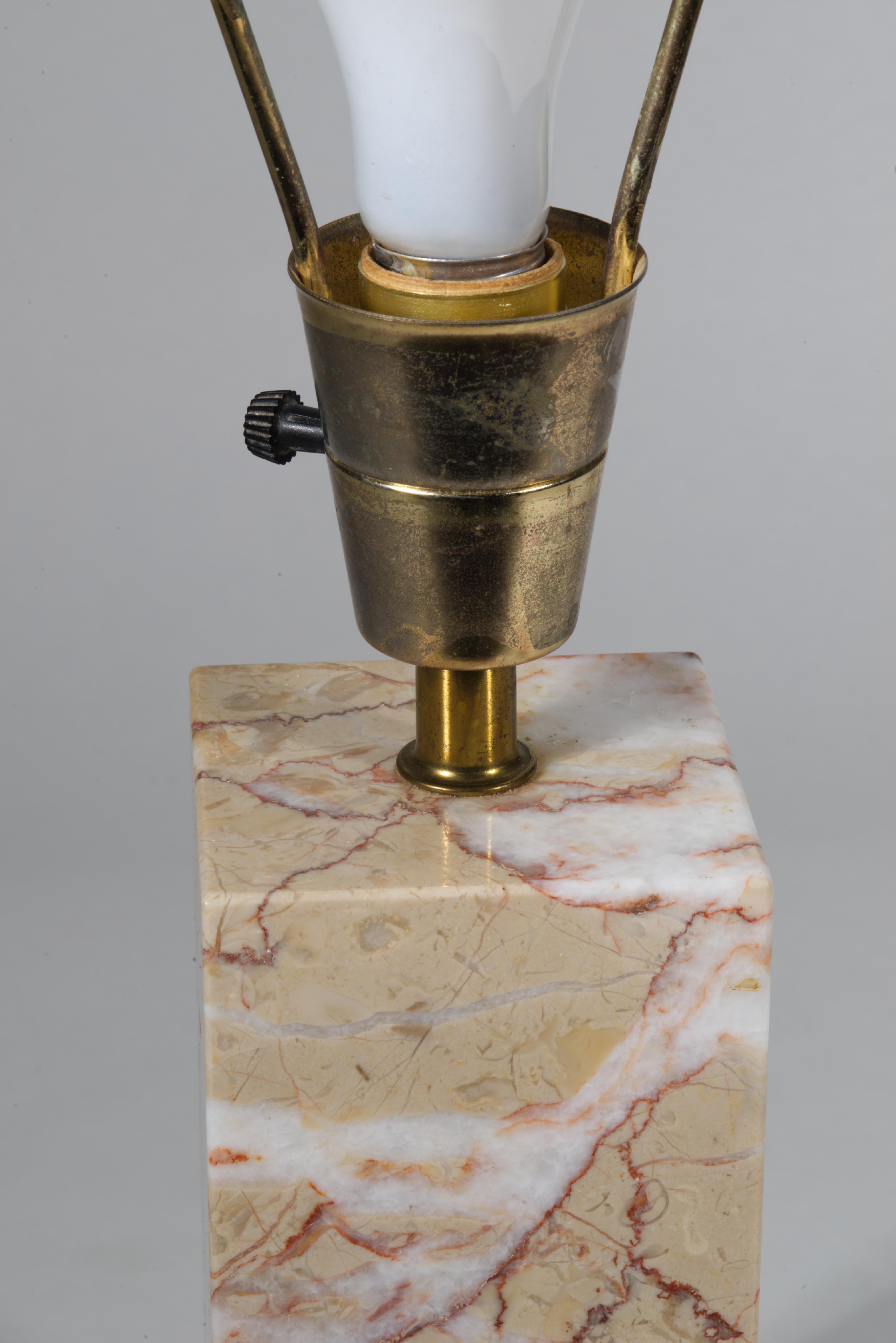 Llamativa lámpara de sobremesa vintage con una alargada base rectangular de mármol, rematada con elegantes apliques de latón. El mármol tiene un gran veteado con colores blanco, marrón y rosa sobre un fondo beige suave. El mármol parece ser Breccia