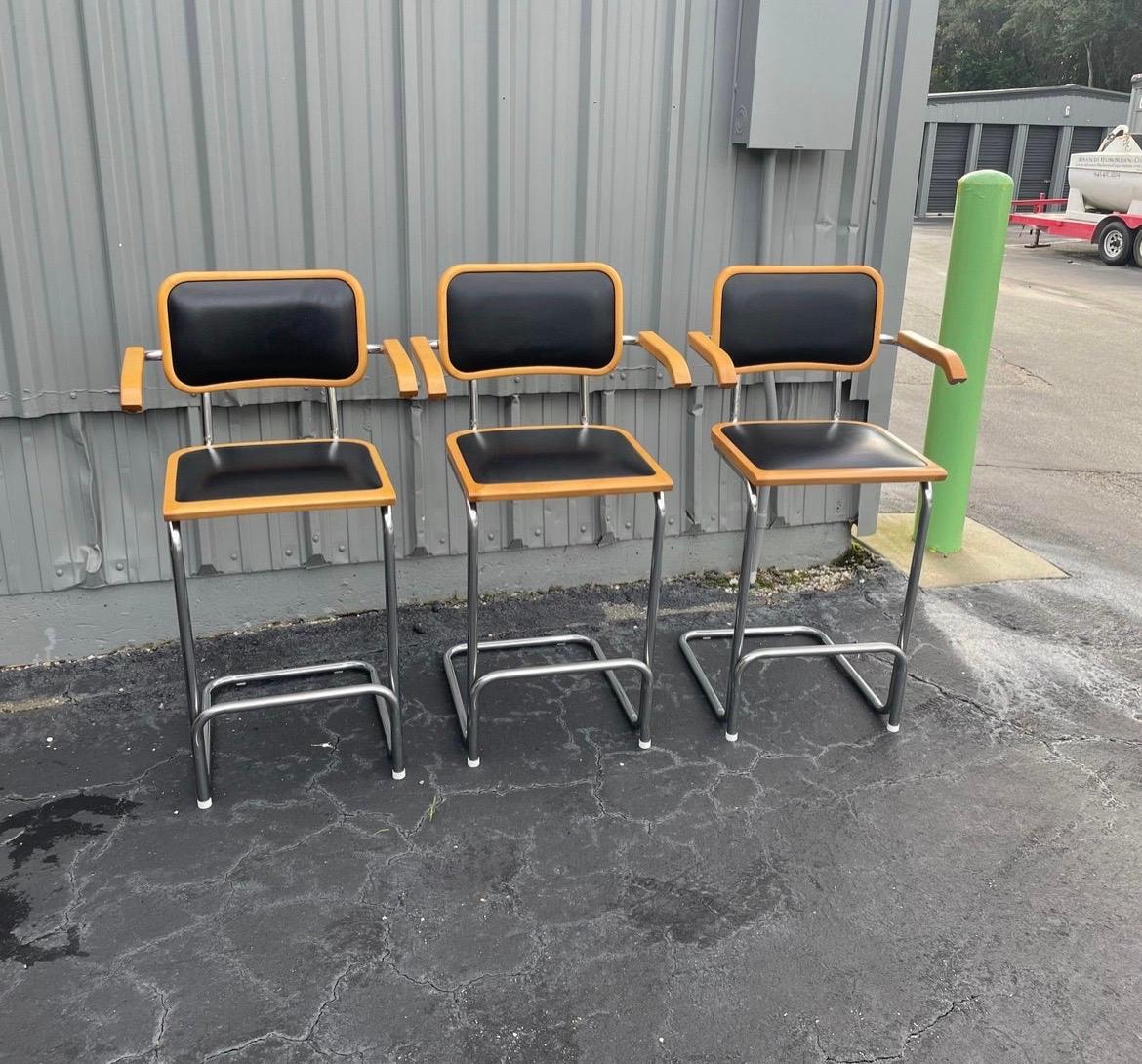 Set of three Marcel Breuer style Cantilever bar stools with black vinyl upholstery. Made by Falcon Products Inc. with the labels still attached. There is also a Made in Italy sticker on two of the chairs. 

Chairs are in overall good condition, with