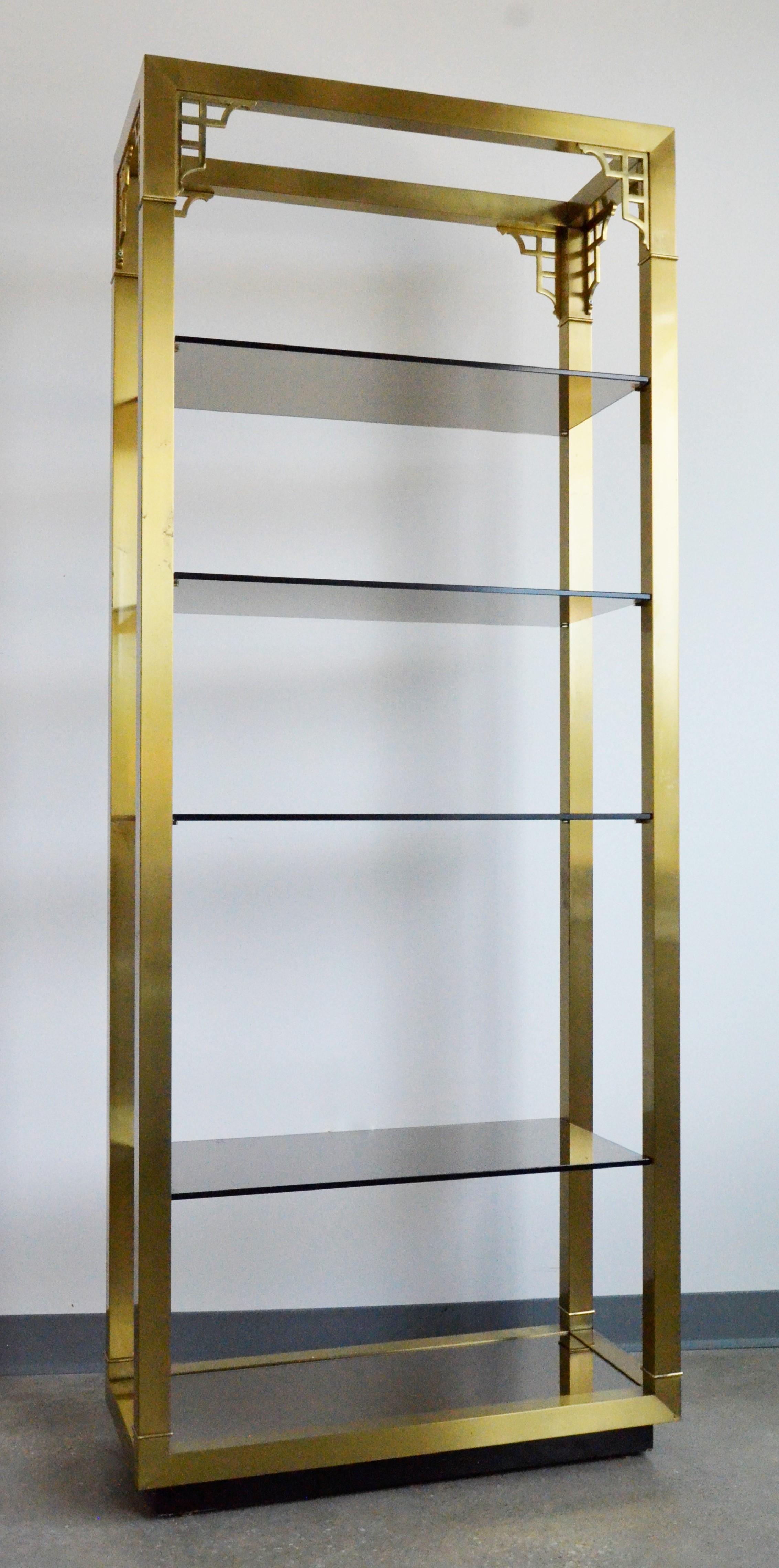 Offered is a Mid-Century Modern Mastercraft style chinoiserie style brass frame with four smoked glass shelves and one mirror bottom shelf étagère. This Hollywood glamour fashion forward piece is jewelry for any room. The étagère has plenty of