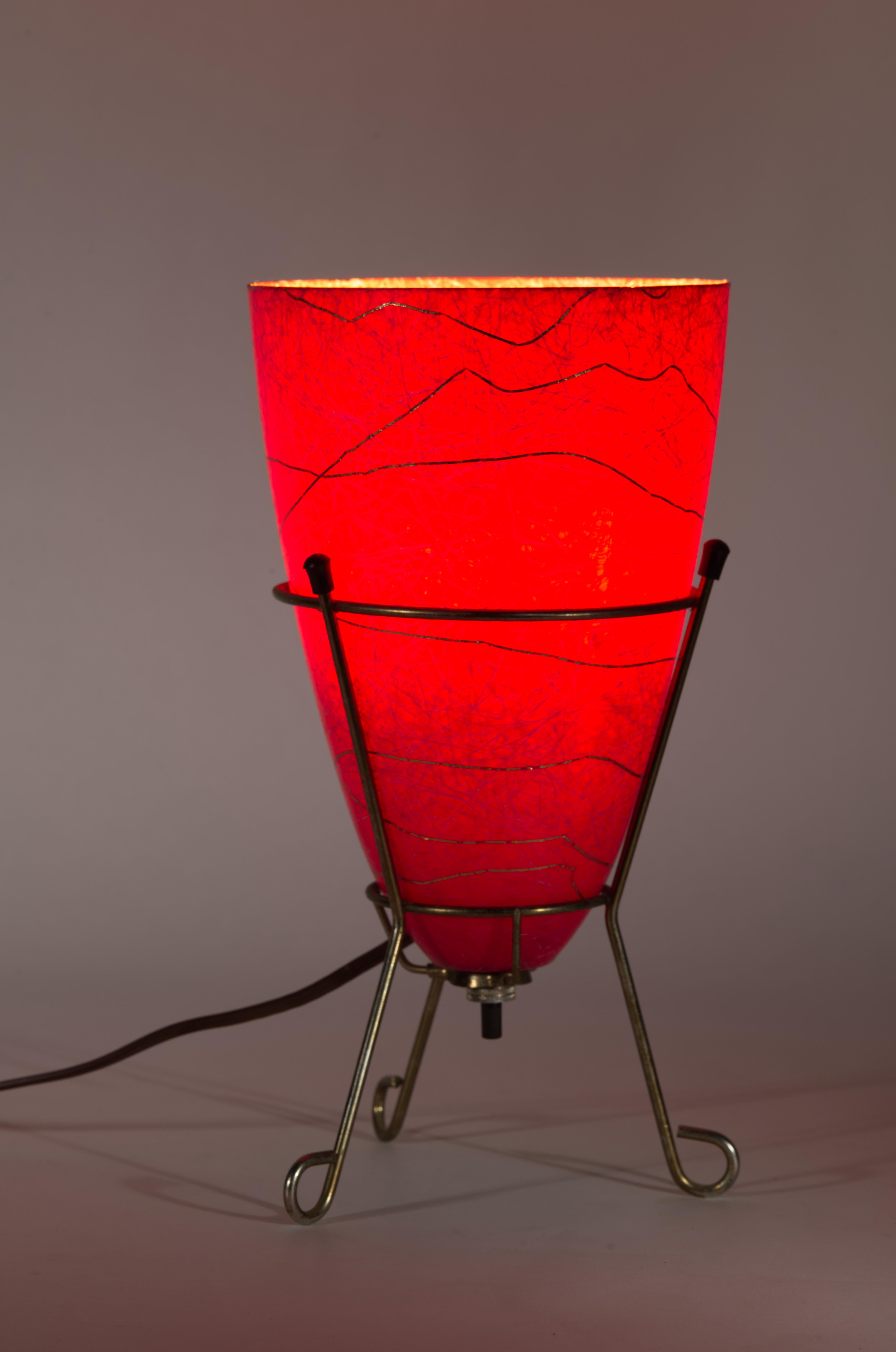 La lámpara de sobremesa o de acento vintage tiene una base trípode de estilo Atomic Modern en metal color latón y una pantalla cónica roscada de fibra de vidrio en rojo con hilos blancos y latón. La forma de la lámpara recuerda a las lámparas de
