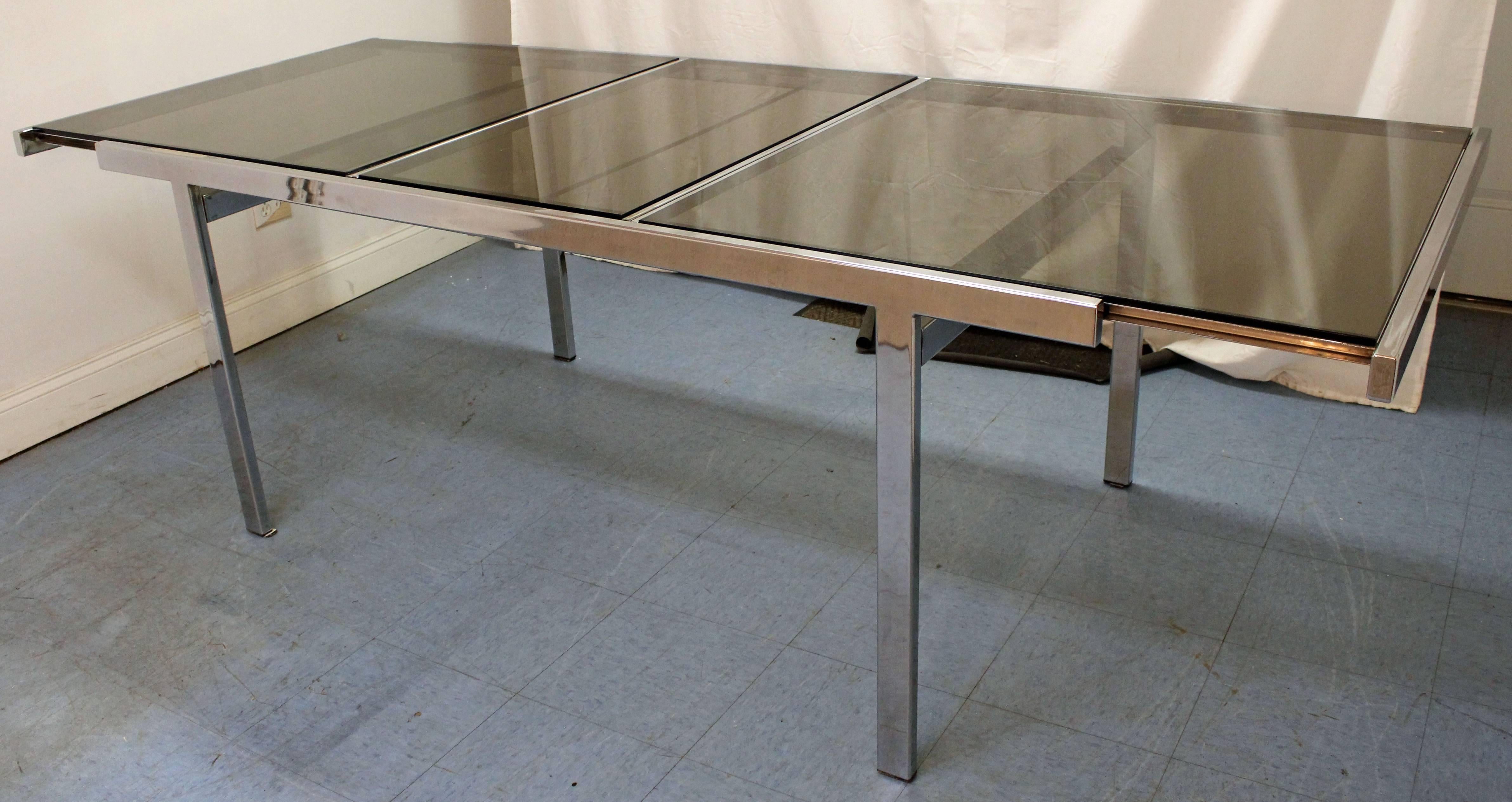 Offered is an extendable chrome dining table, designed by Milo Baughman for Design Institute of America. It is made of chrome with a glass top and features a hidden table leaf that is stored inside the table. See our other listings for a matching