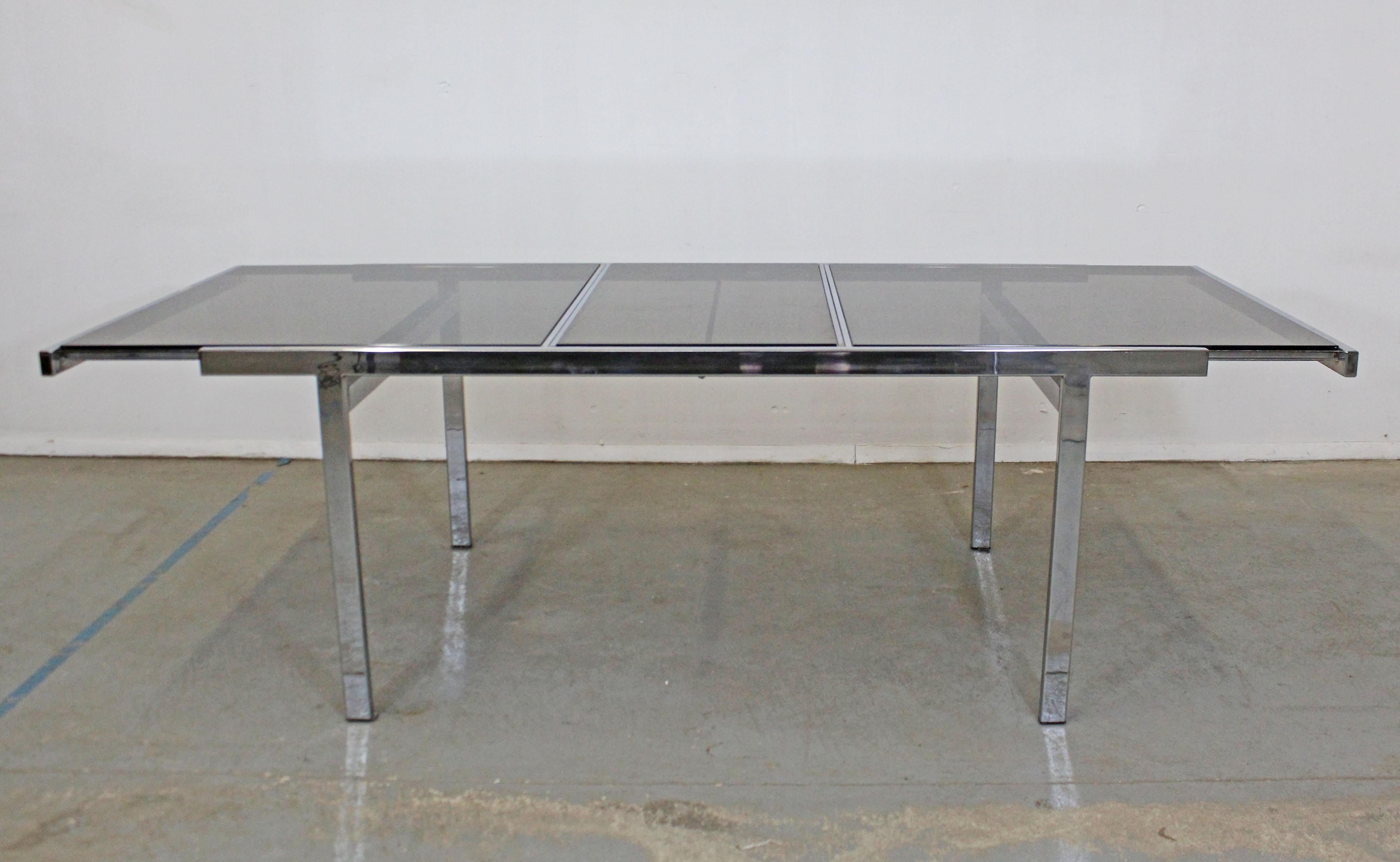 What a find. Offered is an extendable chrome dining table attributed tp Milo Baughman for Design Institute of America (DIA). It is made of chrome with a  smokey black glass top and features a hidden table leaf that is stored inside the table. The