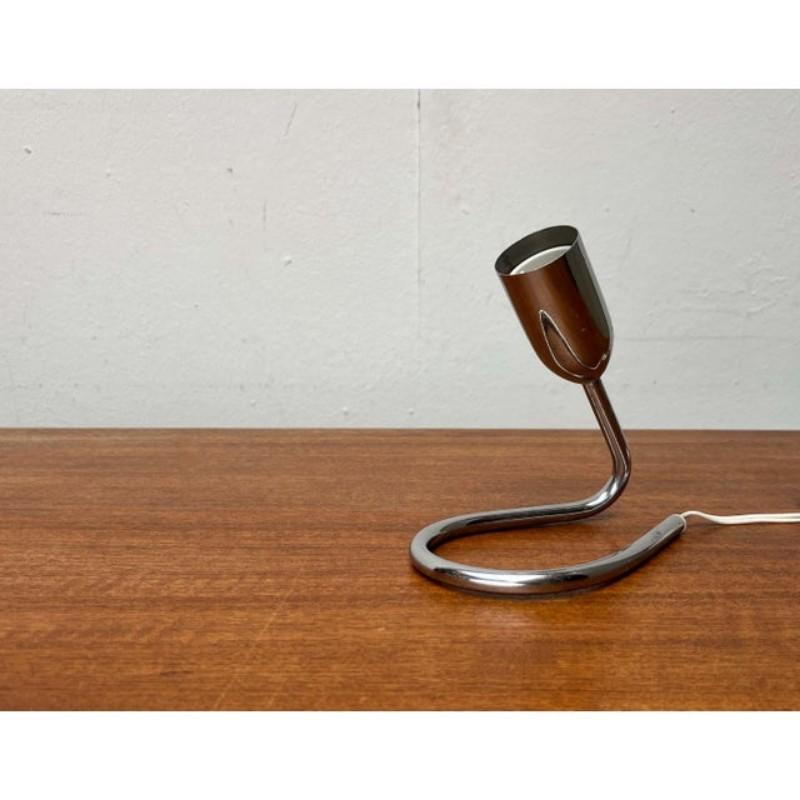 Mid-century table lamp with a striking base.

This small table lamp impresses with its functional simplicity. A chrome-colored metal tube supports the lamp socket at one end. At the other end, it forms a base with a large loop. The cable is hidden
