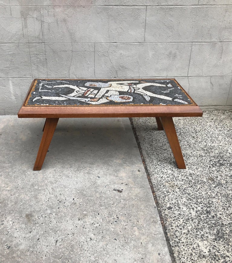A wood and tile top midcentury coffee table, circa 1960s. The tropical hardwood frame and legs with an abstract mosaic tile top showing two dueling figures. Very sturdy, heavy construction.