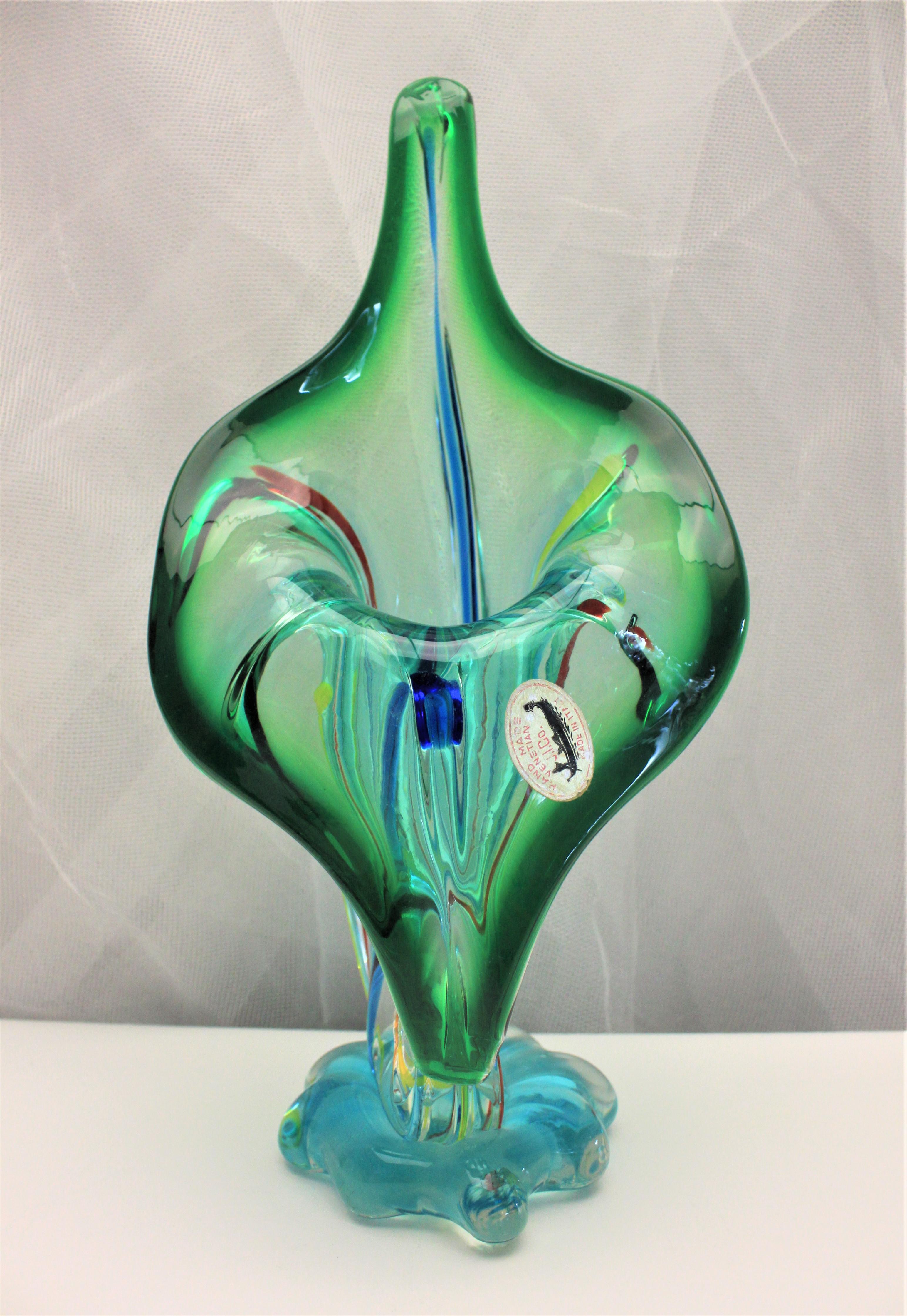 This large jack in the pulpit style art glass Murano vase was made during the 1960s in the Mid-Century Modern style. The vase is done in clear thick glass with a heavy green accent around the opening and with blue, red, and yellow stripes running