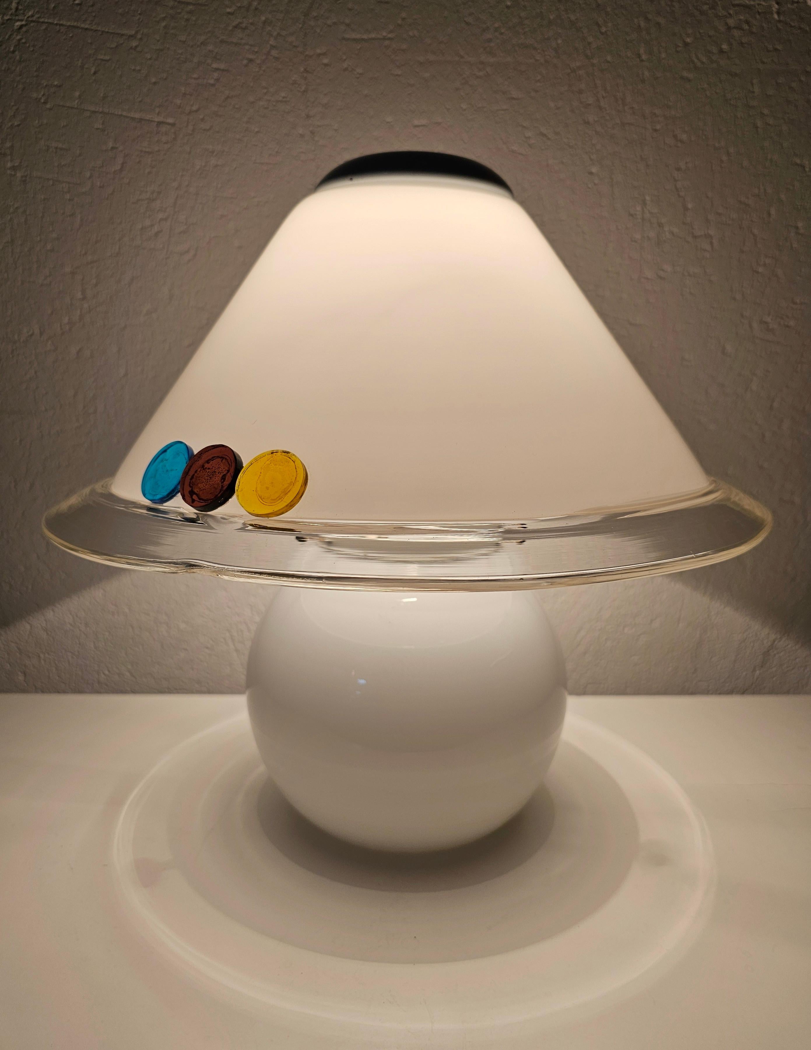 Dans ce listing, vous trouverez une magnifique lampe de table Mid Century Modern en verre Murano blanc avec des détails en verre coloré. Elle est dotée de deux lumières, l'une à l'intérieur de la base de la lampe et la seconde sous l'abat-jour, qui