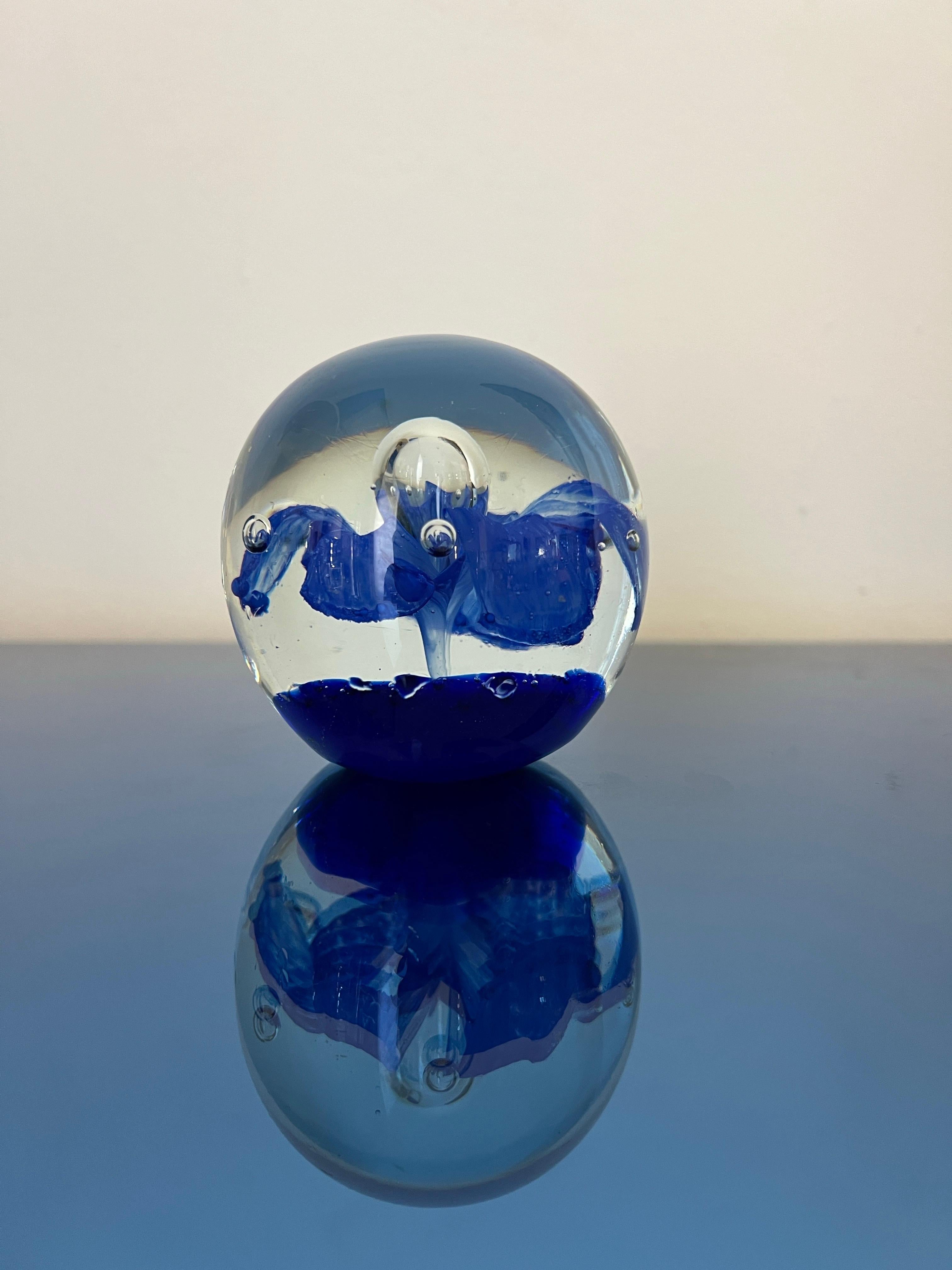 Dieser schöne Briefbeschwerer aus Murano-Glas hat ein einzigartiges Design mit kleinen Luftblasen am Boden, die die zarte blaue Blume im Inneren umgeben. Der Boden besteht aus blauem Glas, in dem die Luftblasen sorgfältig platziert sind, und der