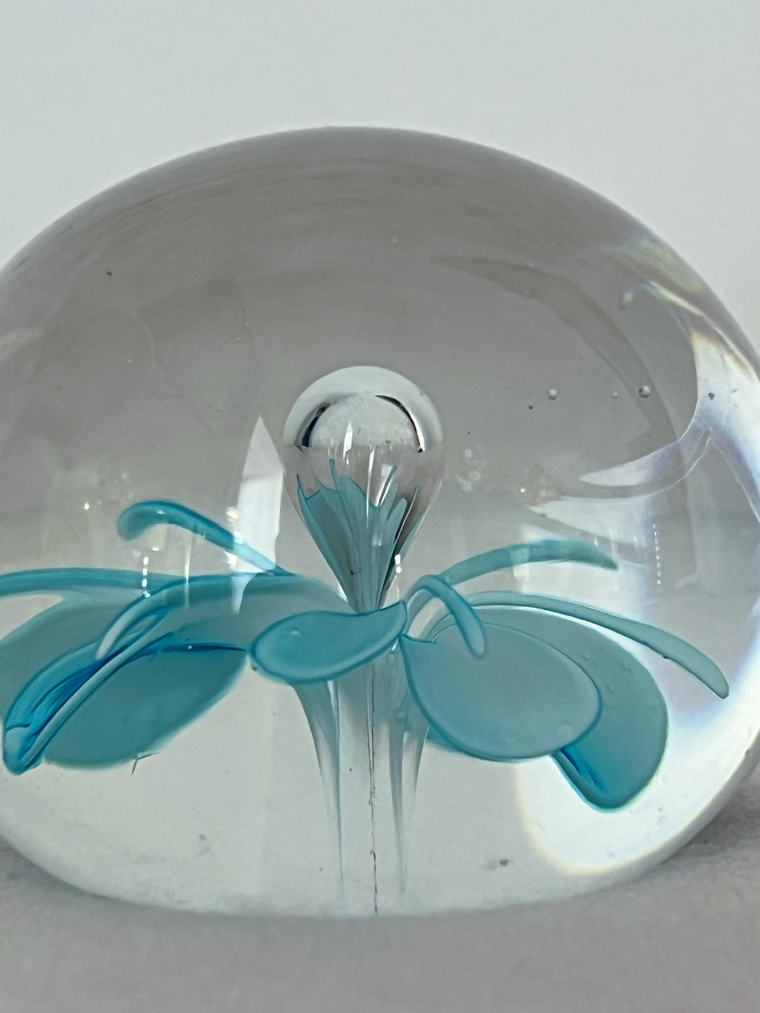 This elegant Murano glass paperweight features a clear, transparent design with a delicate light blue flower inside. Small, subtle air bubbles are suspended within the glass, adding a soft, textured effect that complements the simplicity of the