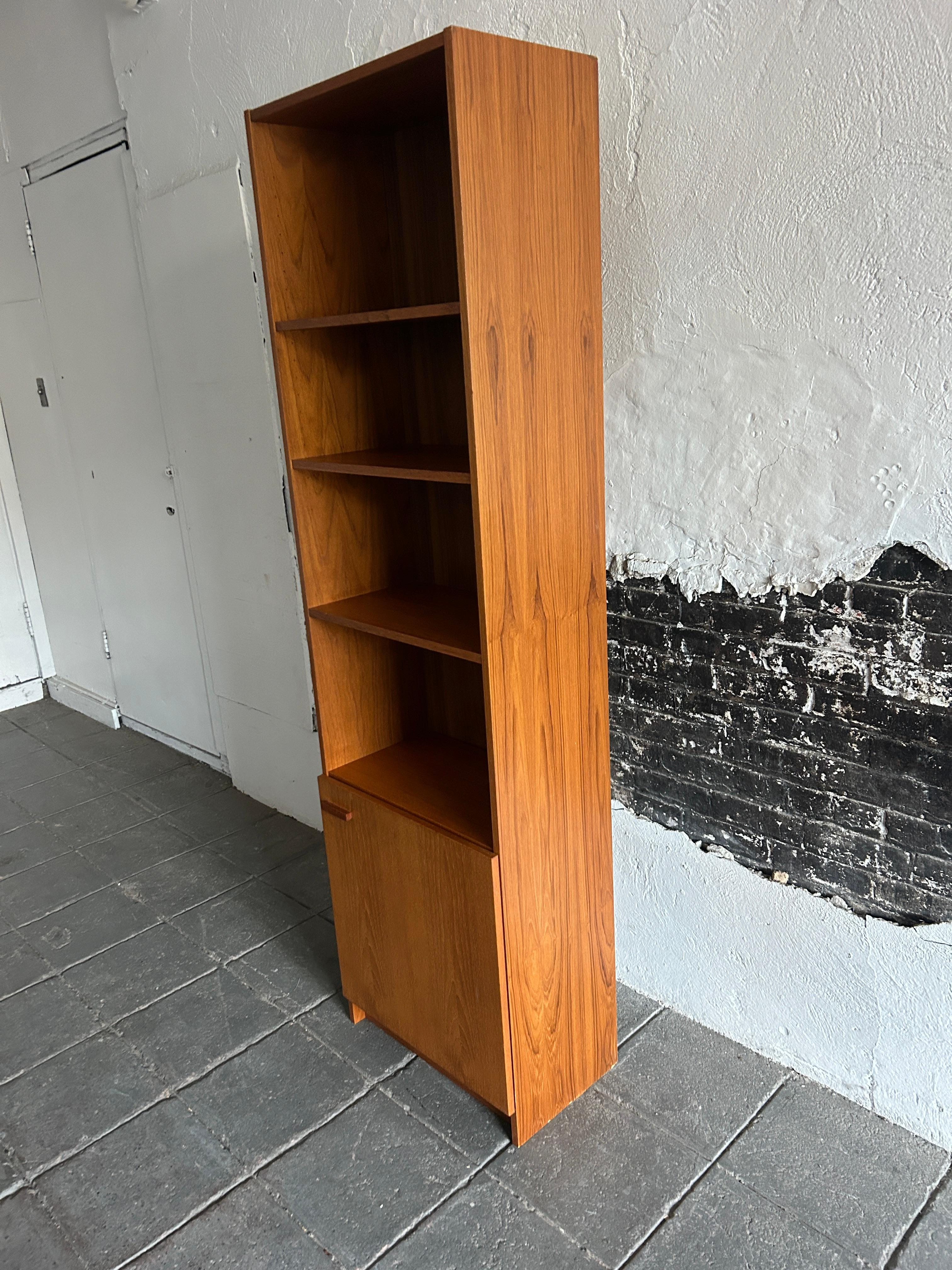 Stunning Mid-Century Modern narrow teak tall bookcase with cabinet door Made in Denmark. This bookcase is completely original and in very good vintage condition. The bookcase has (2) adjustable shelves with 4 shelves total and lower cabinet. The